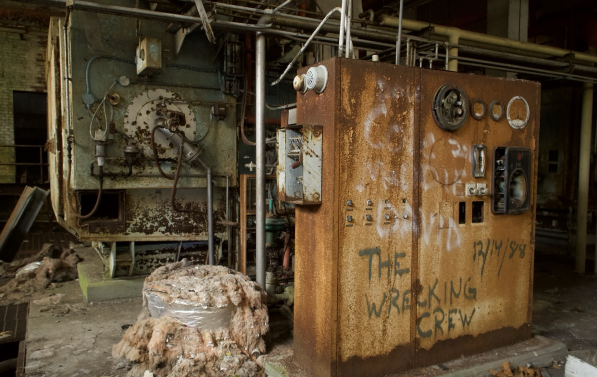 Old rusty industrial machine with gauges and pipes, surrounded by insulation material, in a deteriorated factory setting.