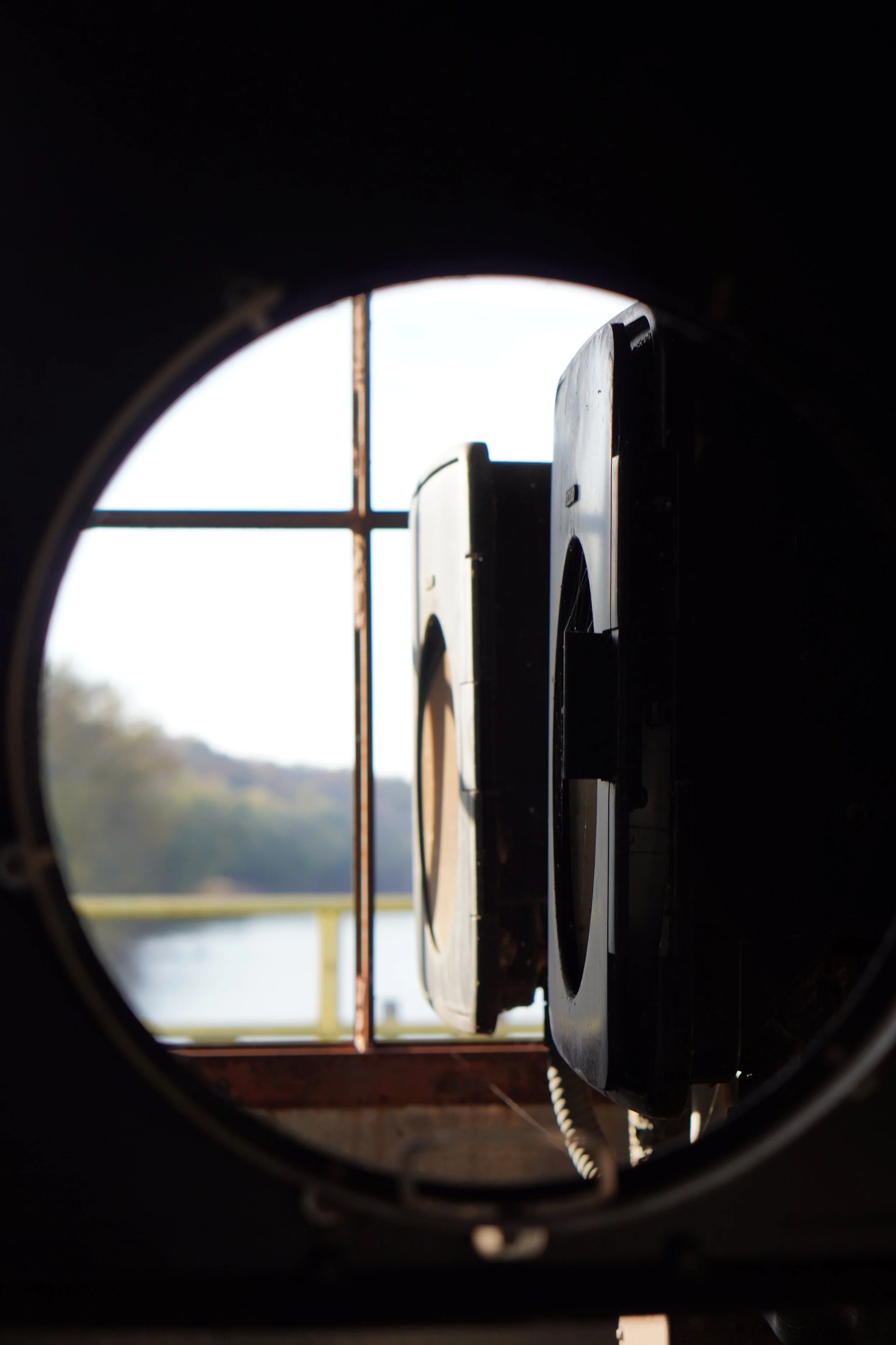 View through a circular opening in a dark structure showing part of a restroom dryer and a large window with a metal grid, with trees and a blue sky outside.