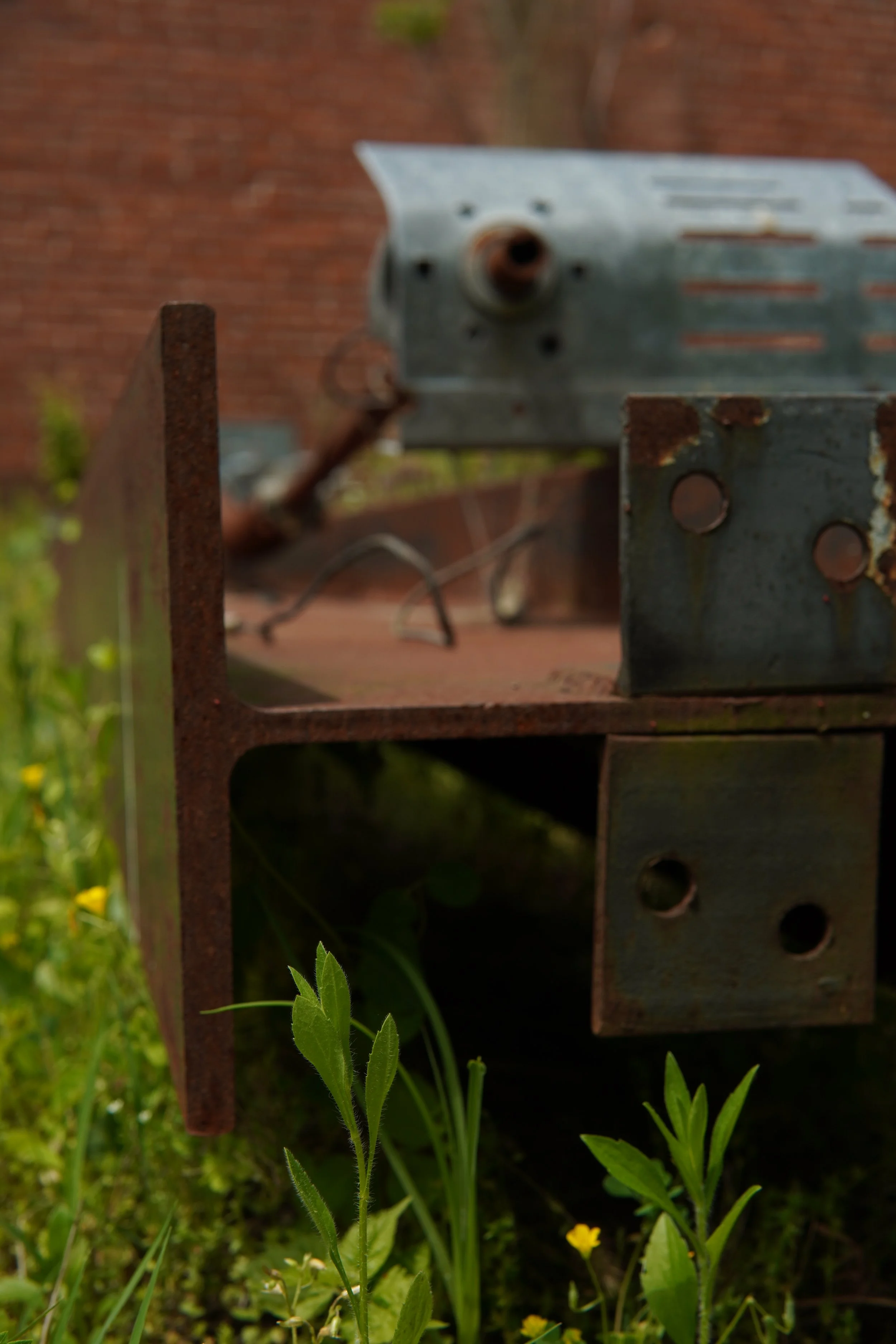 Close-up view of a rusty, old electrical device attached to a metal structure with plants growing at the base. The background shows a brick wall.