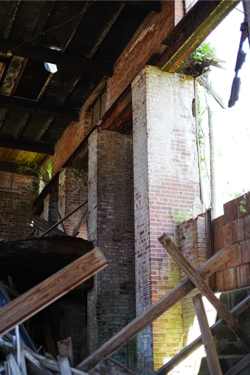Interior view of an abandoned building with exposed brick walls, broken roof panels, scattered wooden beams, and debris on the floor.