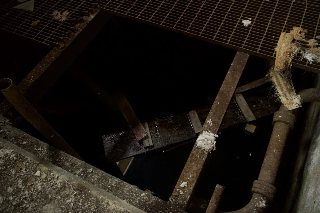 An open manhole cover in a sidewalk, exposing a dark hole with surrounding rusted metal bars, pipes, and concrete debris.