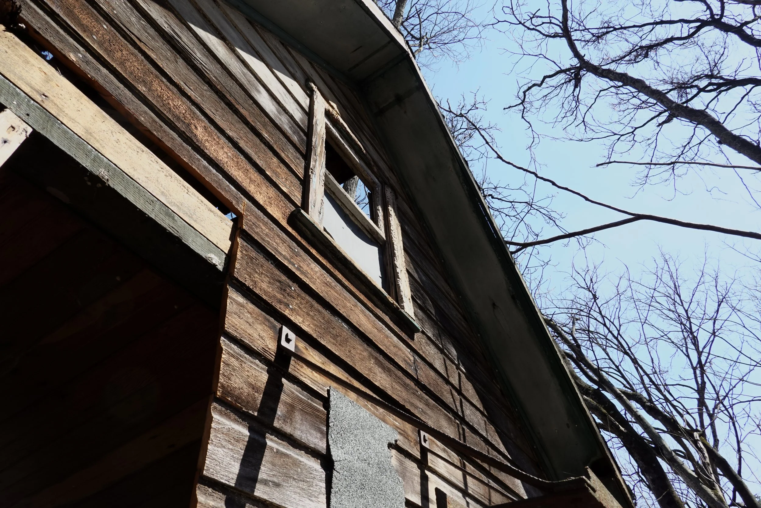 Close-up of an old, weathered wooden house exterior with painted window frames and leafless trees in the background during daytime.