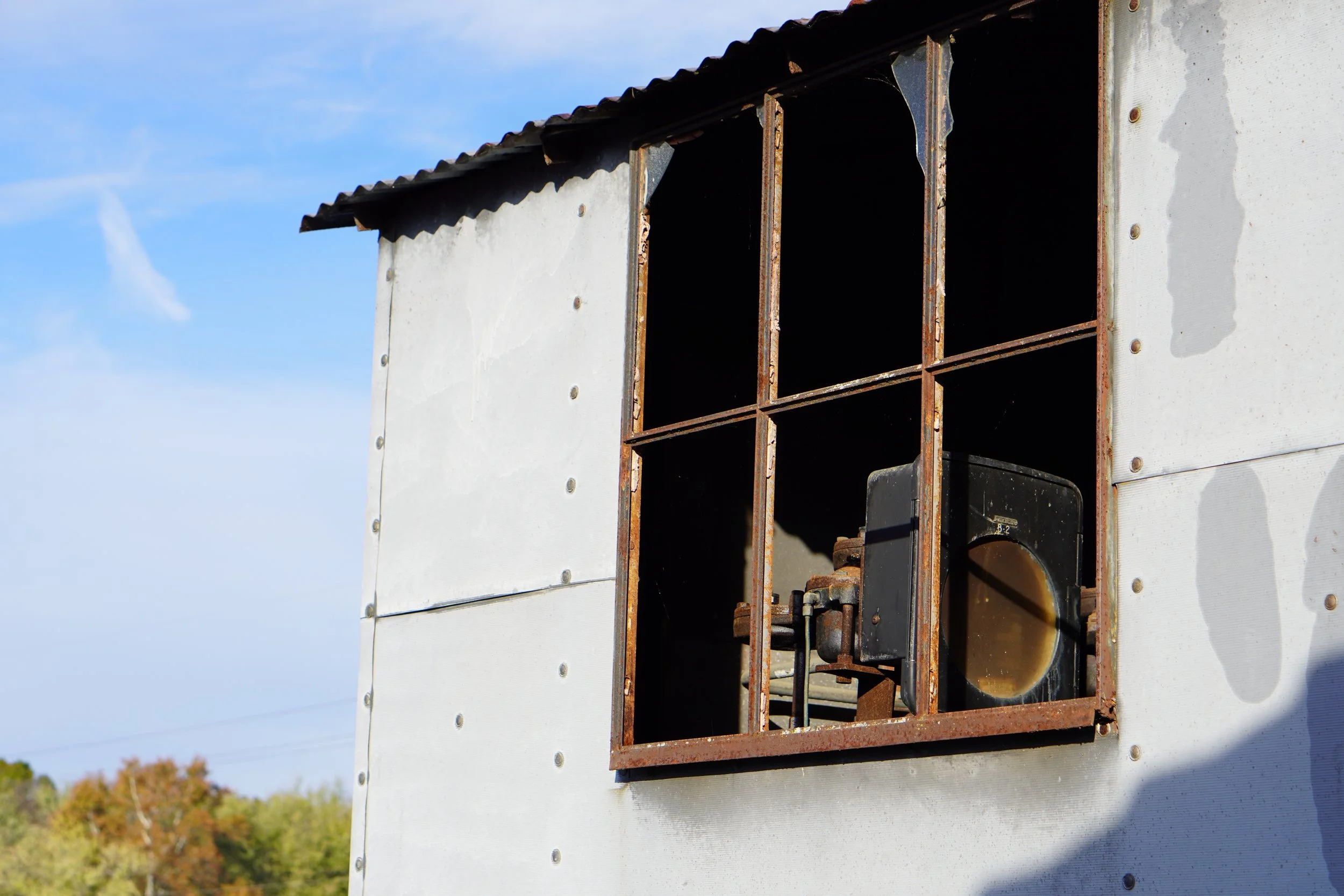 View of a weathered, rusted electrical or utility box with an open, empty metal frame on a white wall, against a blue sky with trees in the distance.