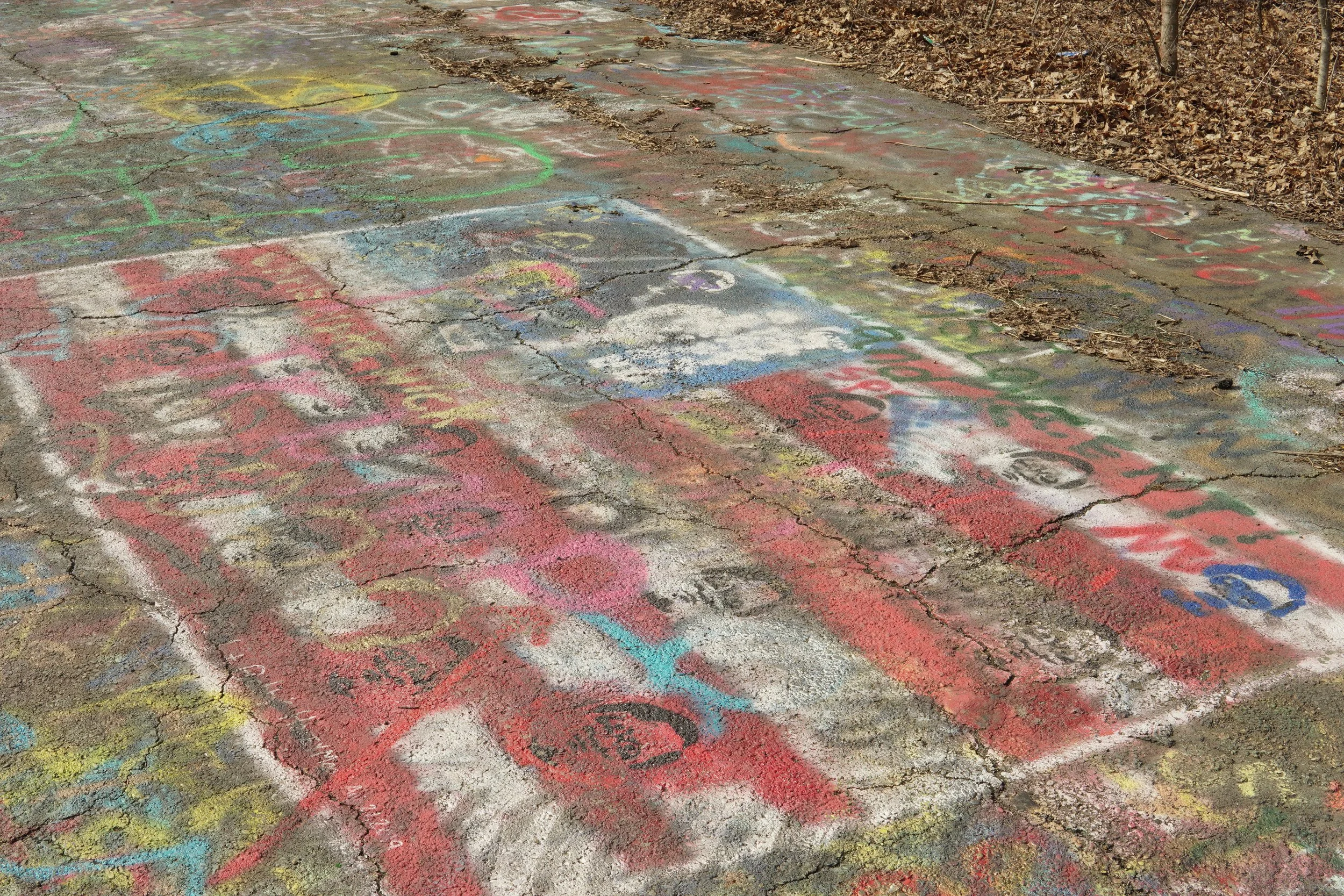 Colored chalk drawings and writings on a cracked concrete sidewalk surrounded by dried leaves and dirt.