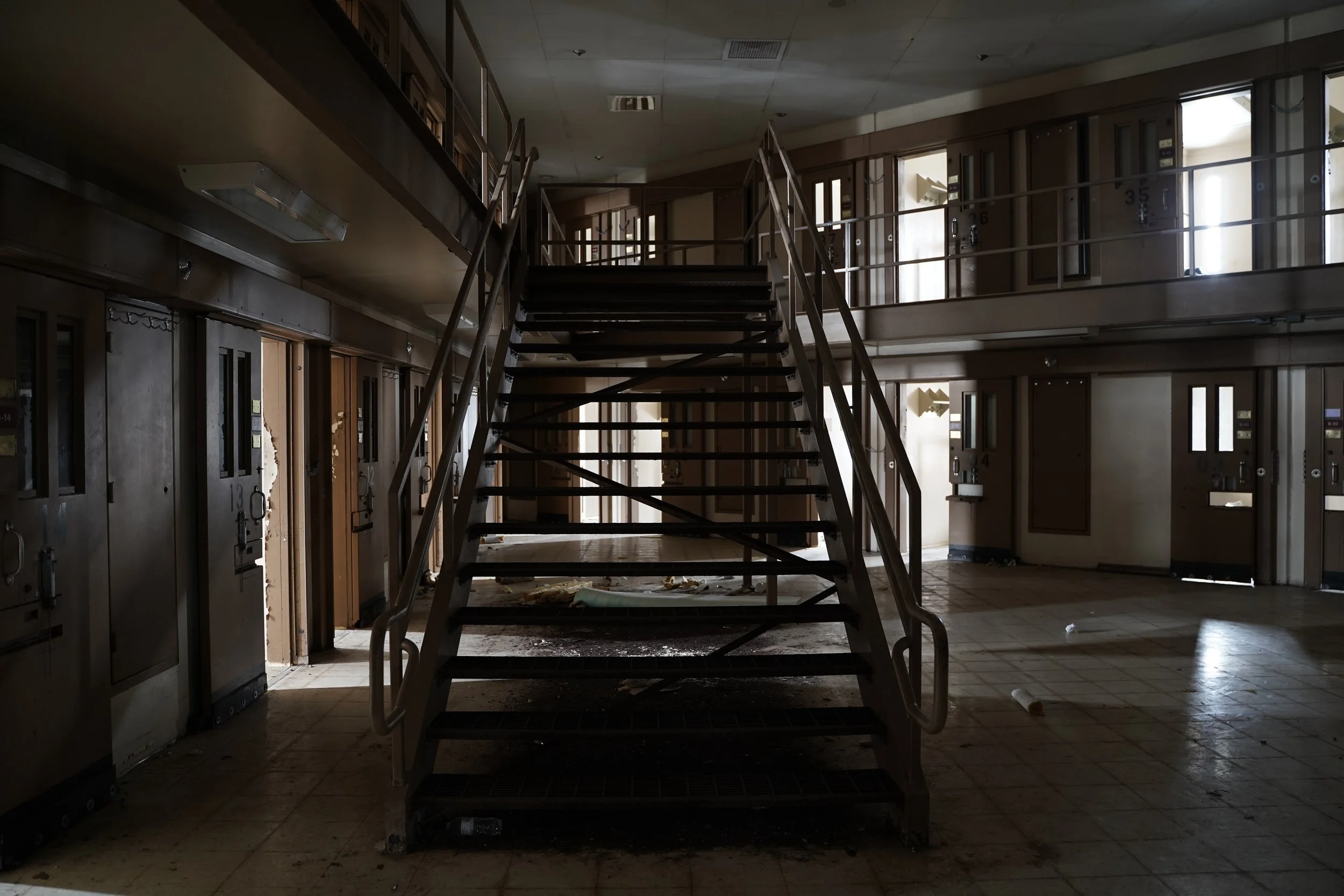 An abandoned, dark interior of a building with a central metal staircase, old lockers on the sides, debris on the floor, and no visible people.