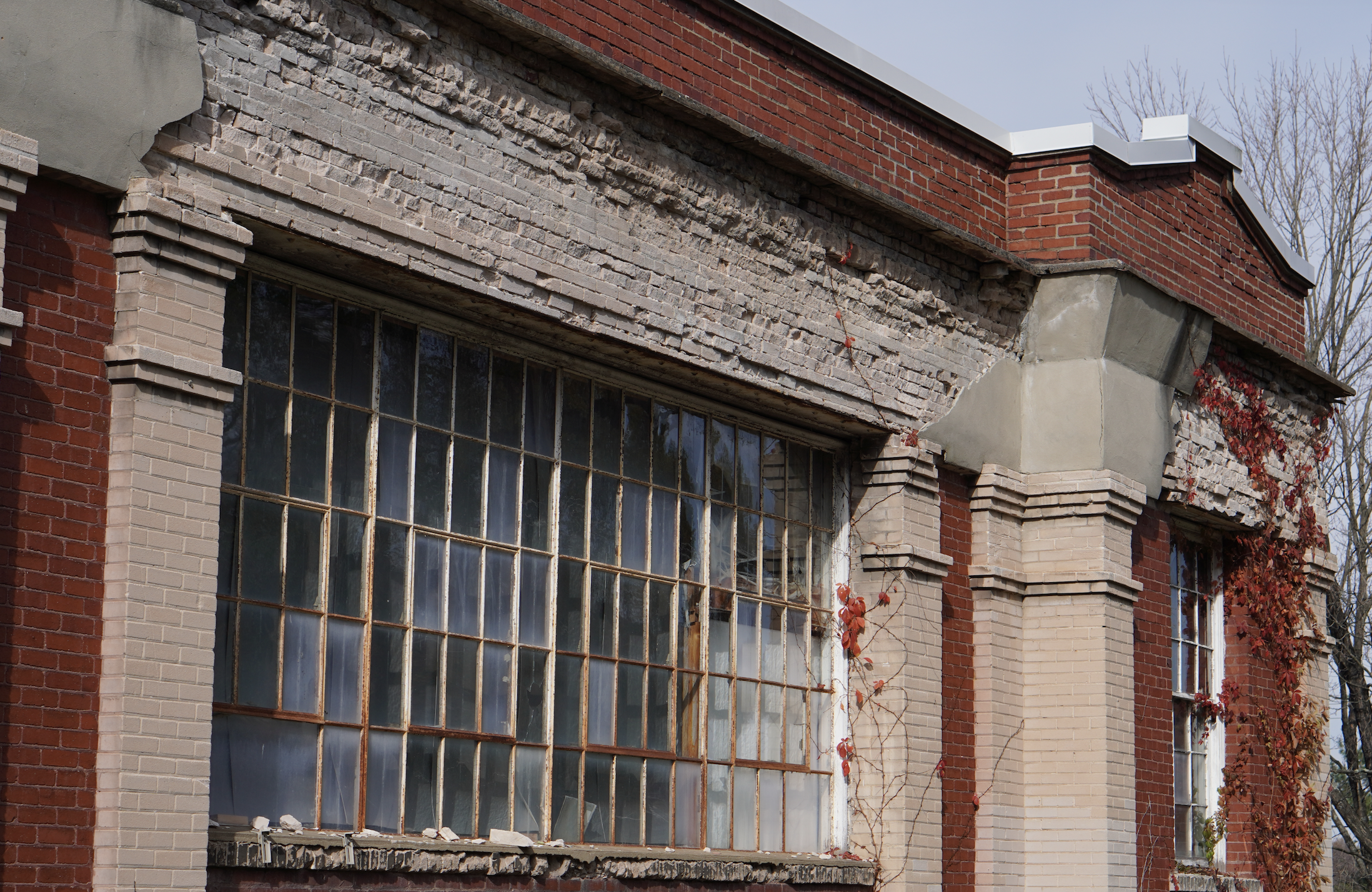 Close-up of a building with old, cracked brick and large window panes, showing signs of disrepair with some panes broken or missing. The building has red and beige brick sections with decorative brickwork and a corner of the roof visible against a cloudy sky. Some vines with red leaves are growing on the building.