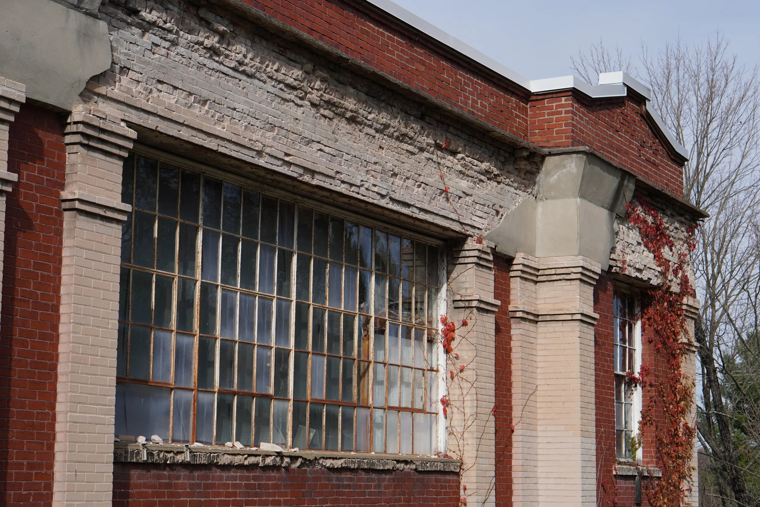 The exterior of an old brick building with large, broken windows and some damaged brickwork, under a clear sky, with leafless trees in the background.