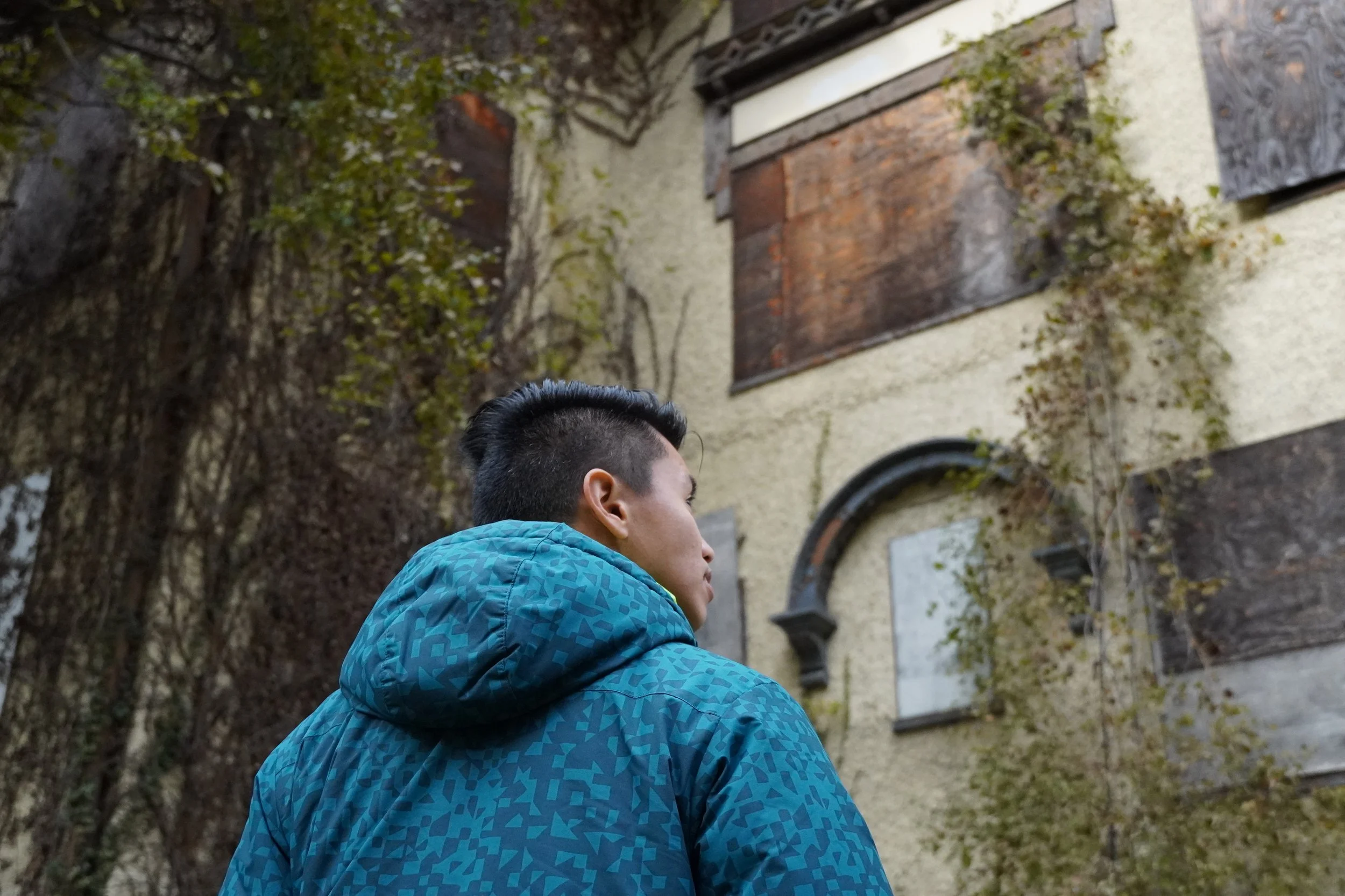A young man with dark hair wearing a blue patterned jacket standing outdoors in front of an old building with boarded-up windows and ivy-covered walls.