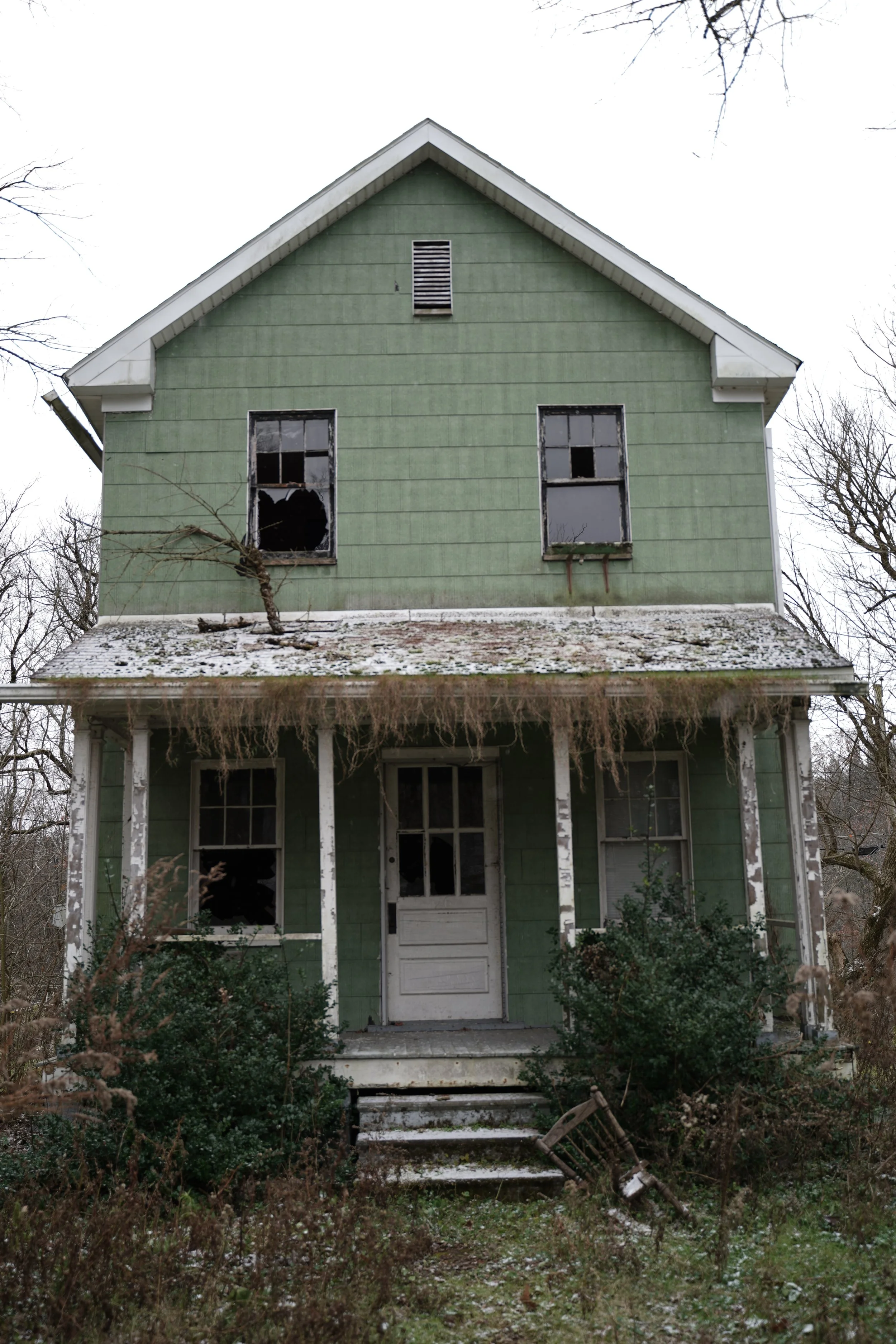 An abandoned, two-story house with green siding, broken windows, and overgrown vegetation. The house has a white front door, a porch with white columns, and a snow-dusted roof.