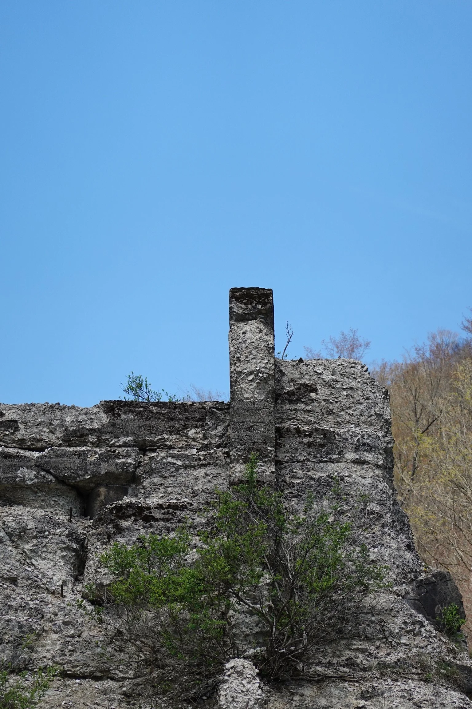 A rocky landscape with ruins of a stone structure and a standing stone chimney under a clear blue sky, with some sparse vegetation and trees in the background.