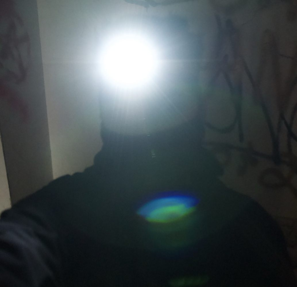 Dark photo of a person taking a selfie with a bright flash, obscuring their face, with graffiti on the wall in the background.