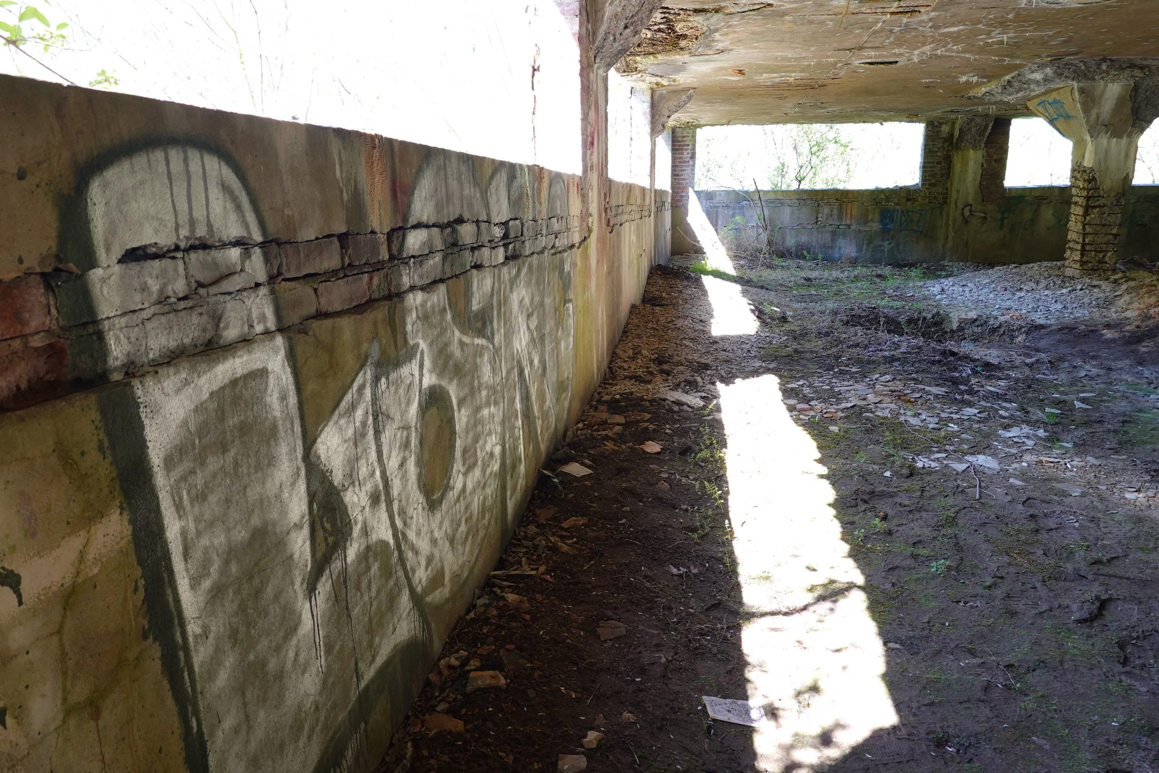 Interior of an abandoned building with brick and concrete walls covered in graffiti, dirt, and debris on the floor, and sunlight streaming in through broken windows.