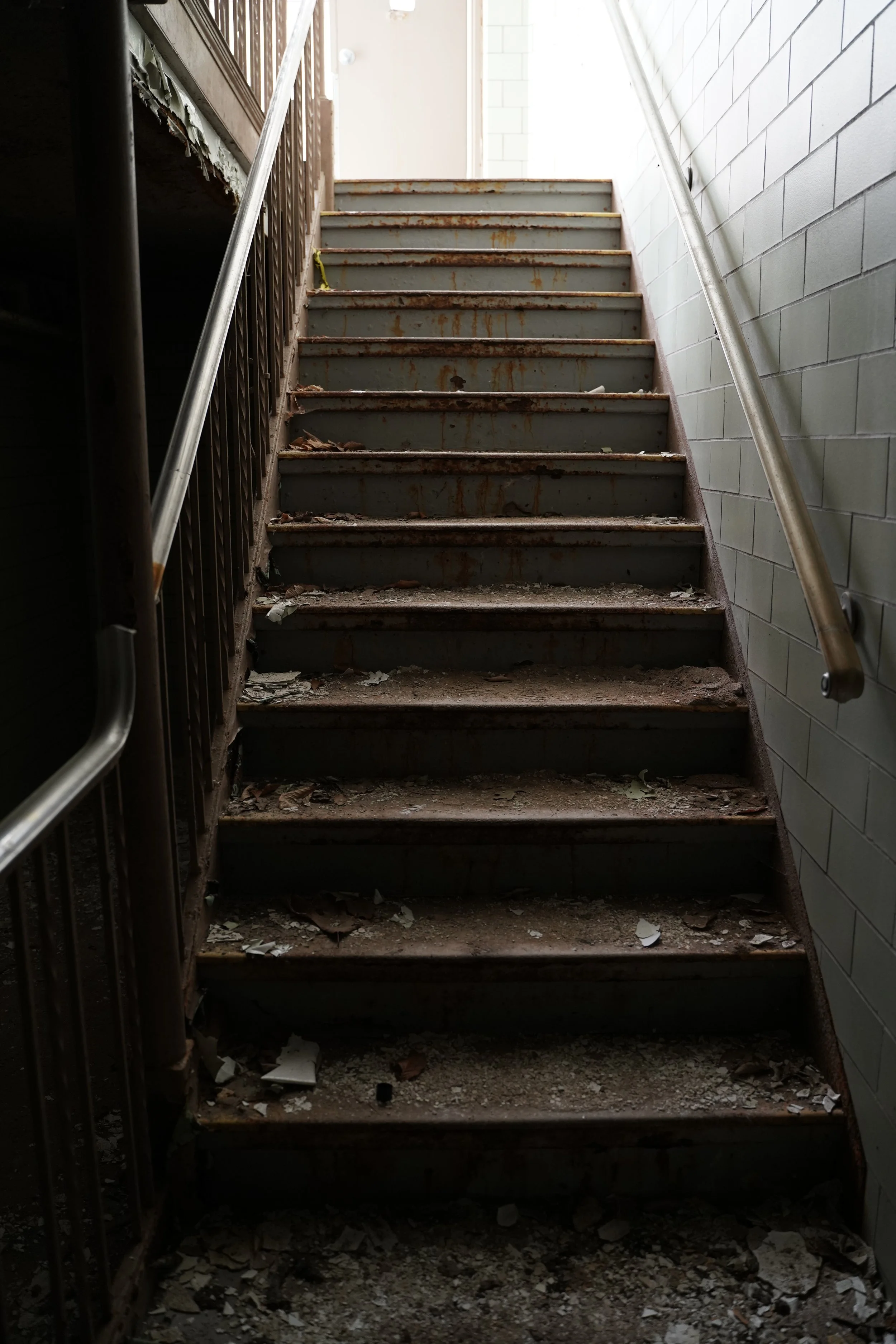 A dusty, rusty staircase with debris and torn paper on the steps, flanked by a metal railing on each side, leading upwards past a white brick wall on the right and an industrial-style brown metal structure on the left.