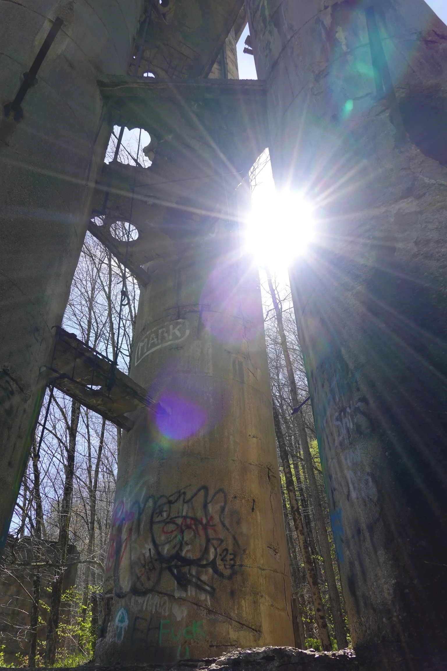Sunshine shining through an abandoned structure with graffiti on concrete pillars and trees in the background.