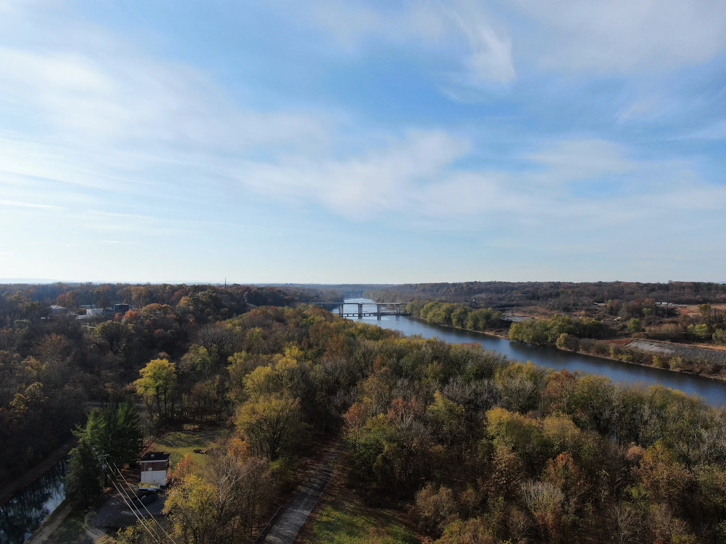 A scenic landscape view of a river winding through a forest with trees showing autumn colors, a bridge crossing the river, and a blue sky with some clouds overhead.