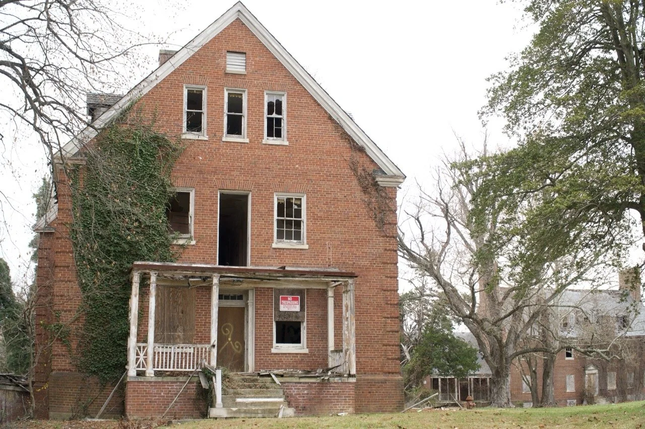 An abandoned, dilapidated brick house with broken windows, overgrown ivy, and a sagging front porch with a 'No Trespassing' sign, surrounded by leafless trees.