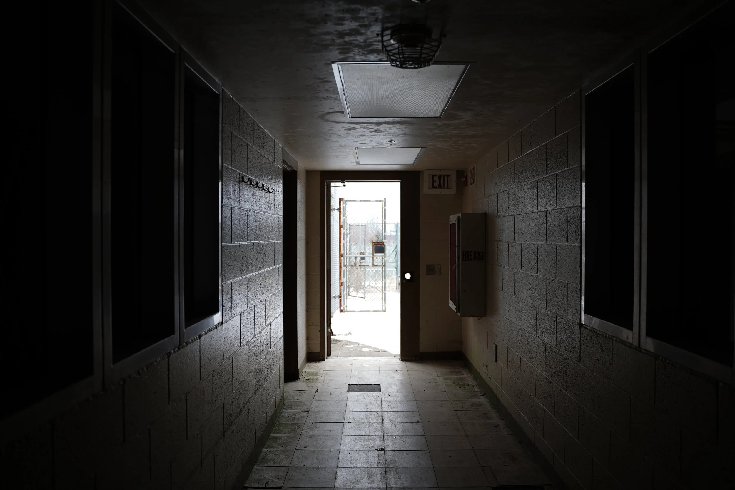 Dark indoor corridor with tiled walls and ceiling, leading to an open door at the end showing a chain-link fence outside, with an exit sign and fire hose box along the wall.