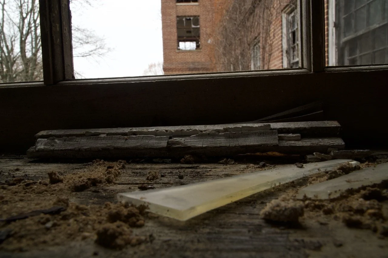 A dusty windowsill with broken wood and debris, looking outside at a brick building with open windows and leafless trees.