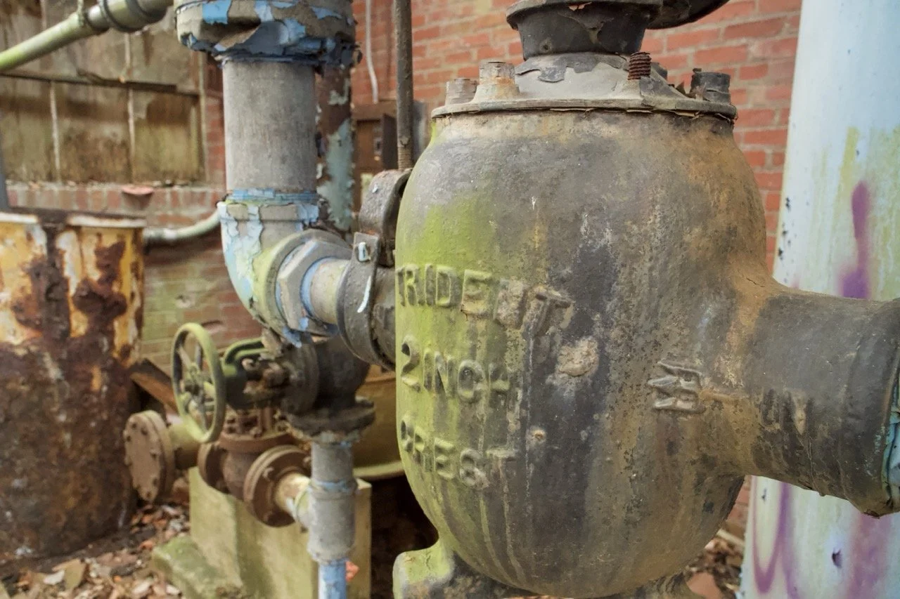 Close-up of an old, rusted water meter with pipes attached, situated outdoors near a brick wall.