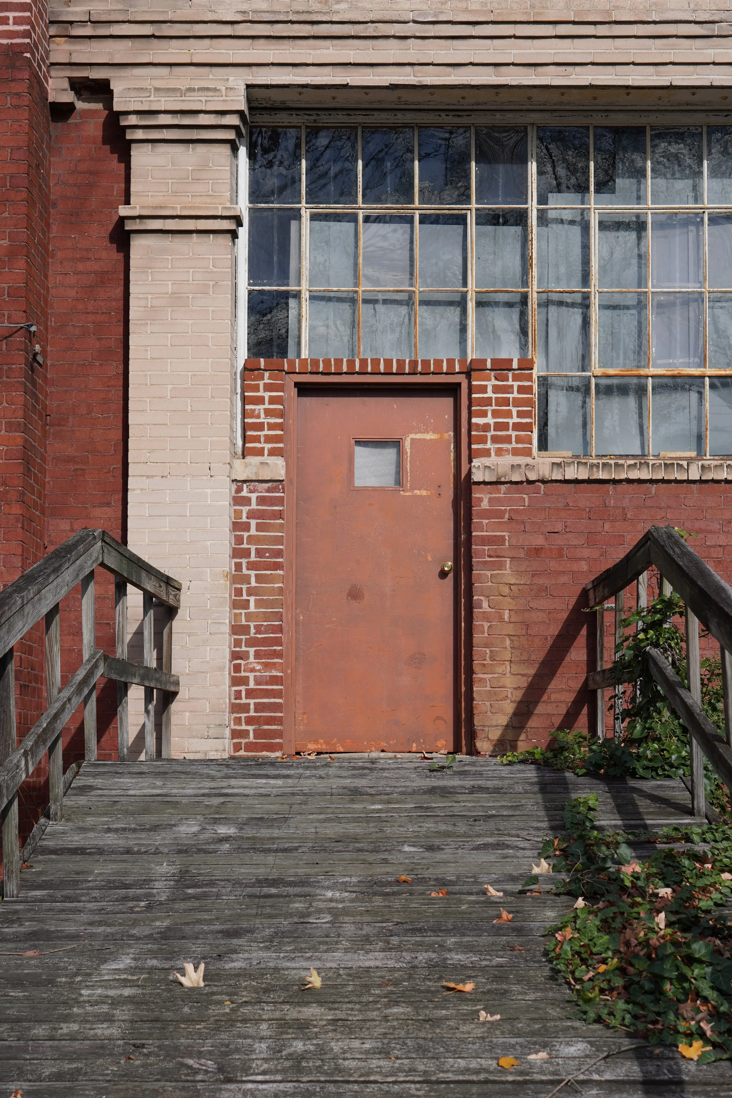 A weathered industrial-style brick building with large windows and a rusted metal door, flanked by wooden railings and overgrown ivy on the steps.