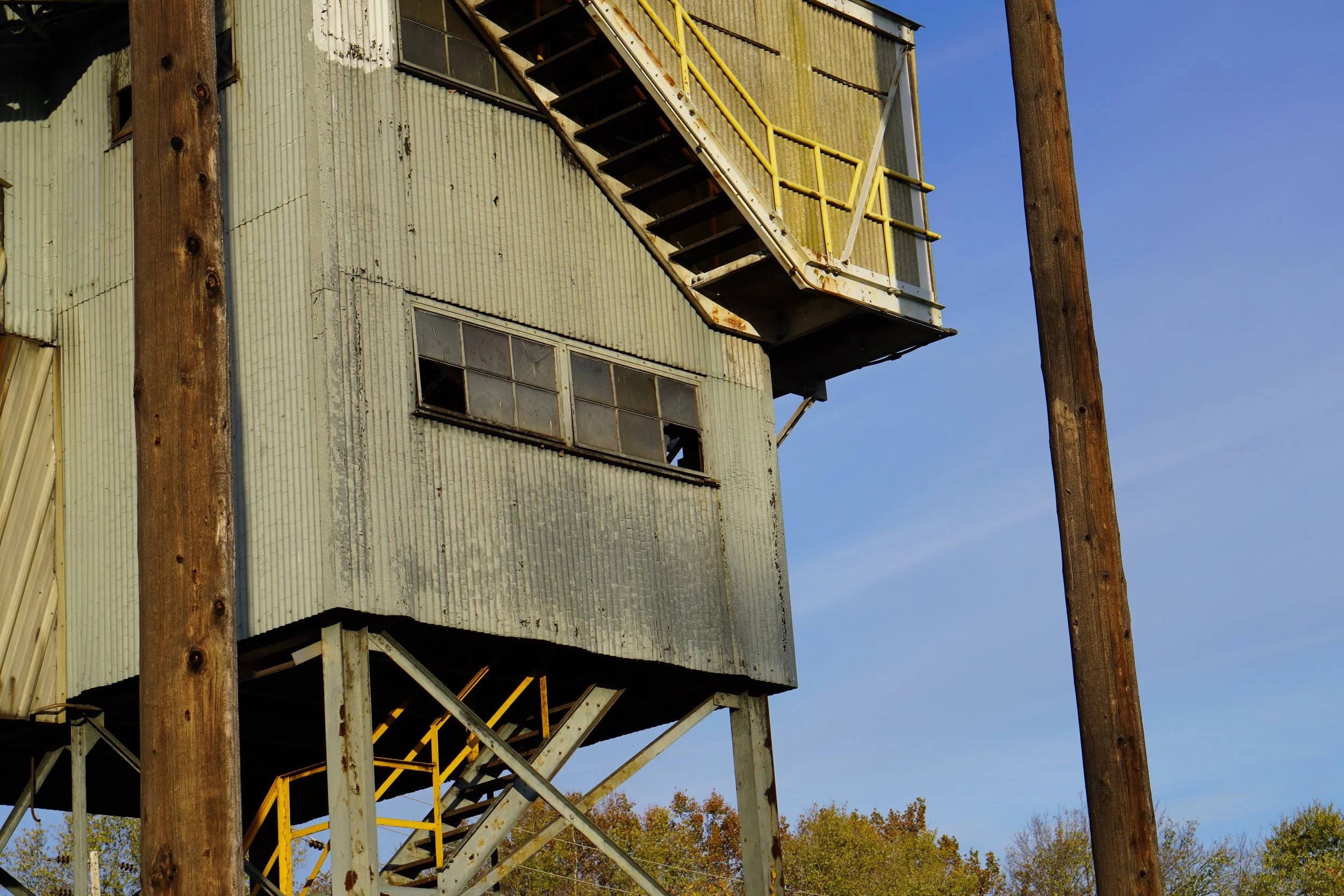 Close-up of an old, weathered building with metal siding, a metal staircase with yellow handrails, and two large wooden utility poles against a clear blue sky.
