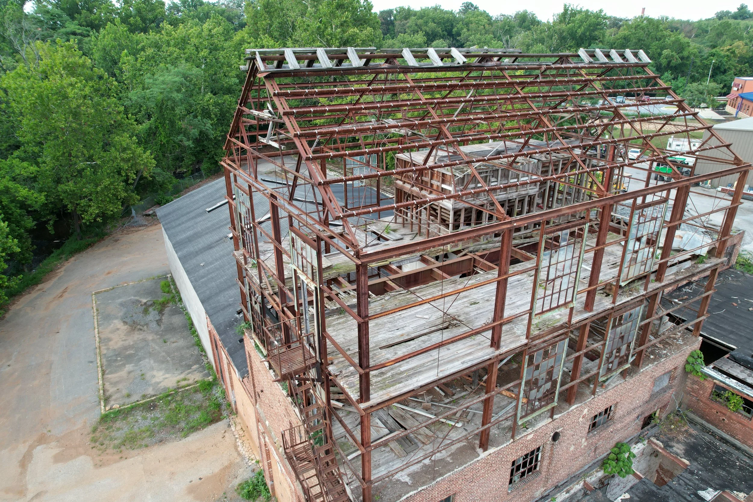 Aerial view of a building under renovation with a wooden roof frame, surrounded by green trees and parking lots.