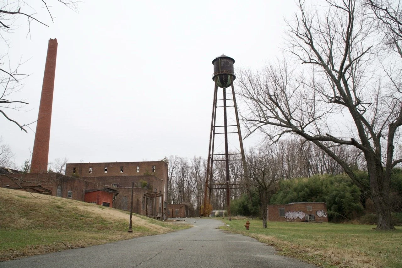 An abandoned industrial site with a tall, rusted water tower, a brick building, a tall smokestack, leafless trees, and graffiti on a smaller building, under an overcast sky.