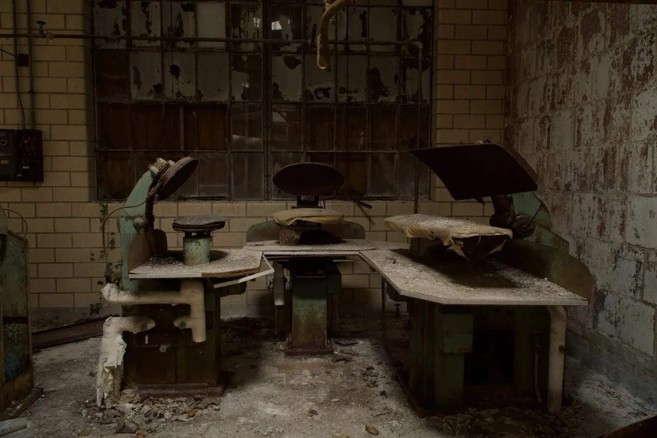 An abandoned, rusty office or industrial workspace with corroded chairs and tables, dirt, and peeling paint on the walls.