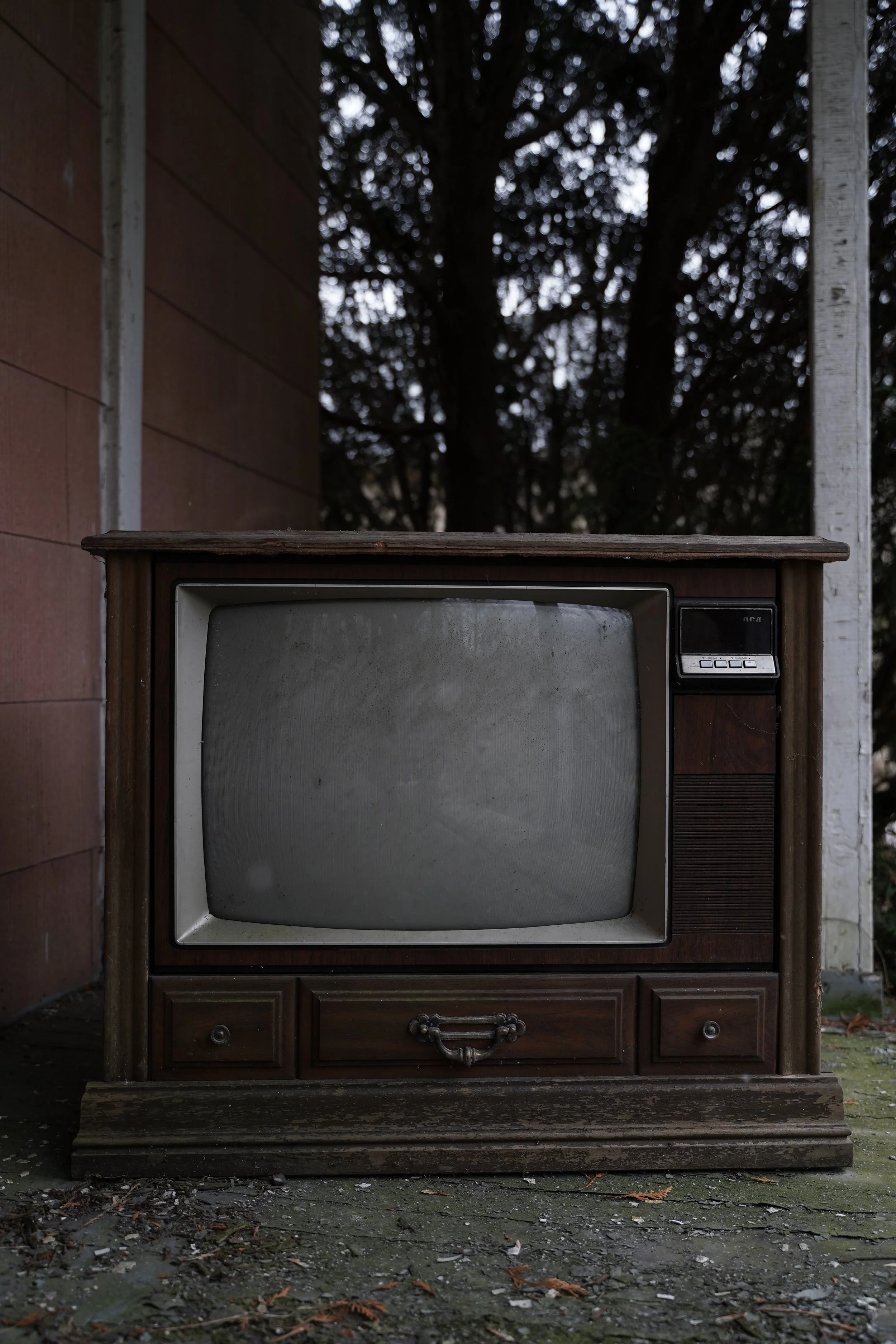 Old, vintage television set outdoors on weathered ground with fallen leaves, against a backdrop of a building wall and dark trees.