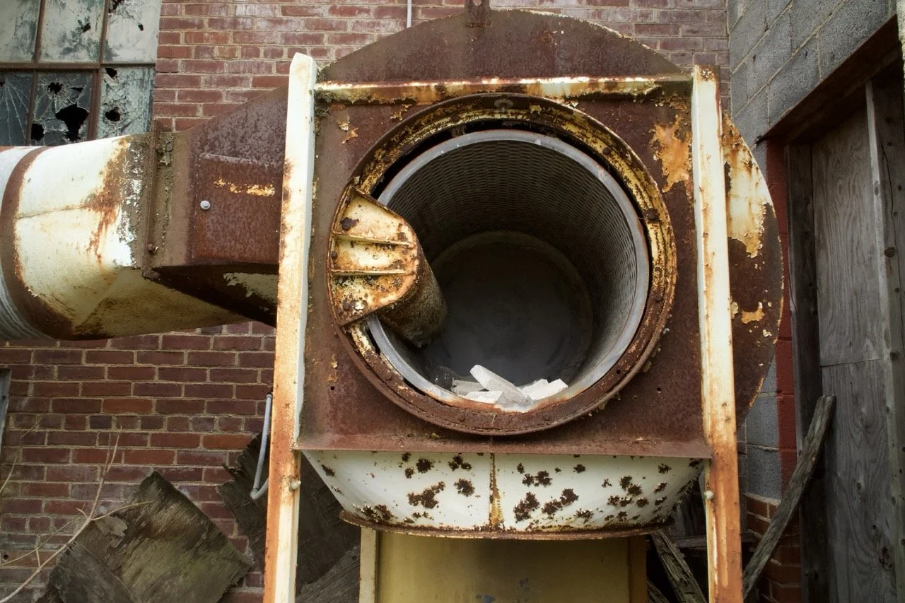 An old, rusty industrial dryer or kiln with a large circular opening, set against a brick wall with broken windows, surrounded by weathered wooden boards and debris.