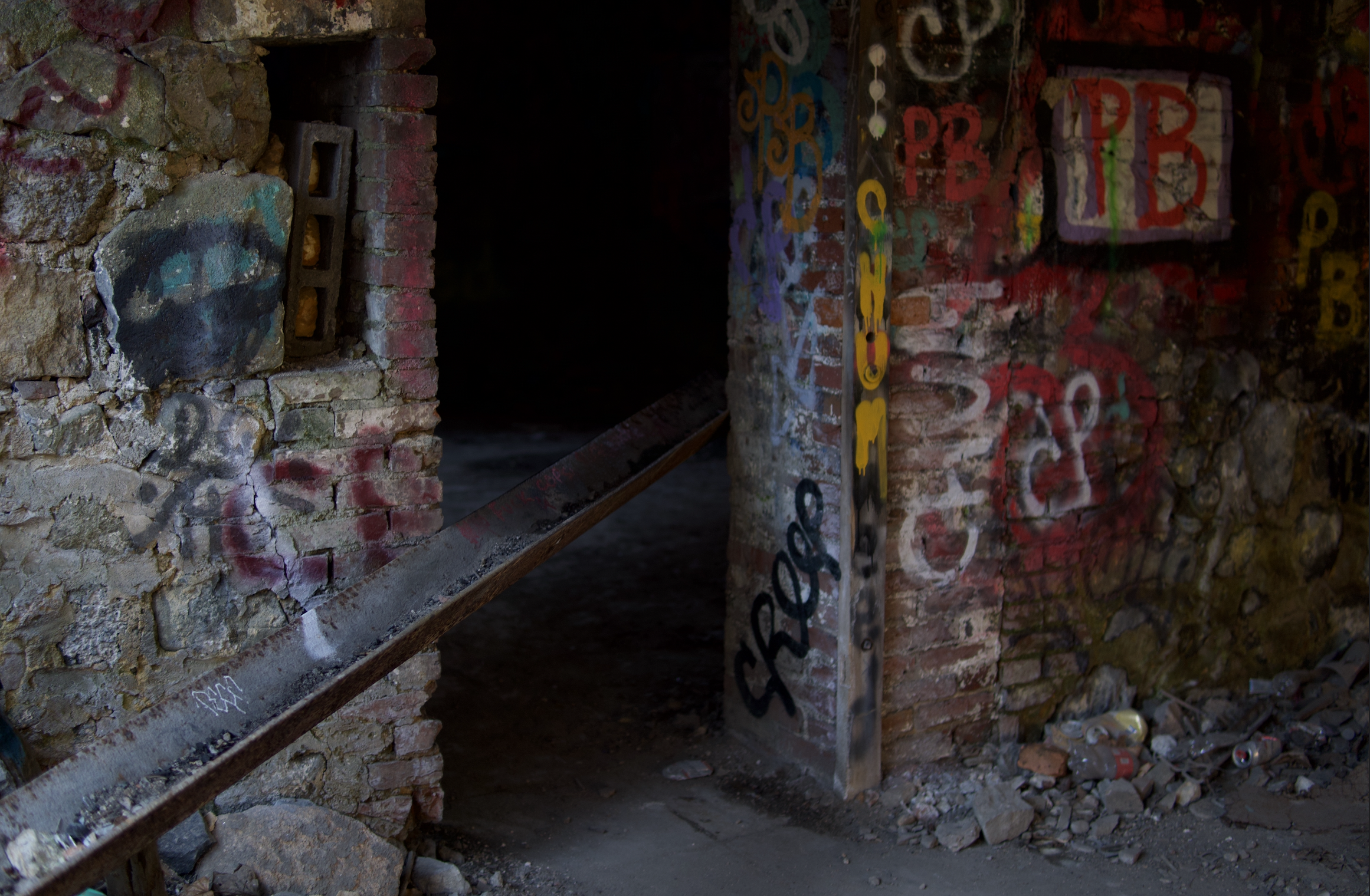 A photo of an abandoned brick structure with graffiti on the walls, including spray-painted letters and designs, and scattered debris on the ground.