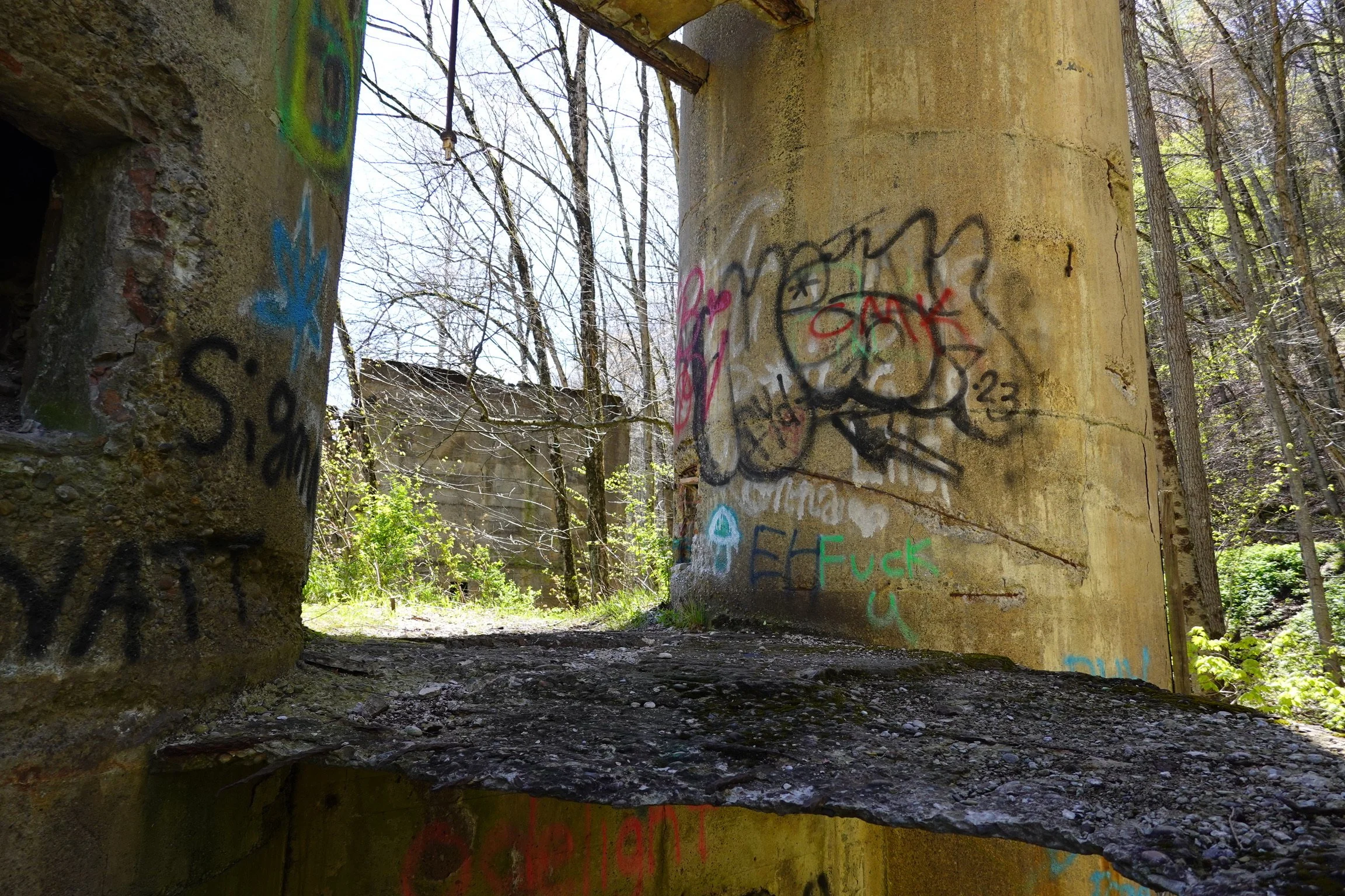 The photo shows a partially destroyed concrete structure in a wooded area with graffiti on its walls. The ground is rocky, and leafless trees are visible in the background.