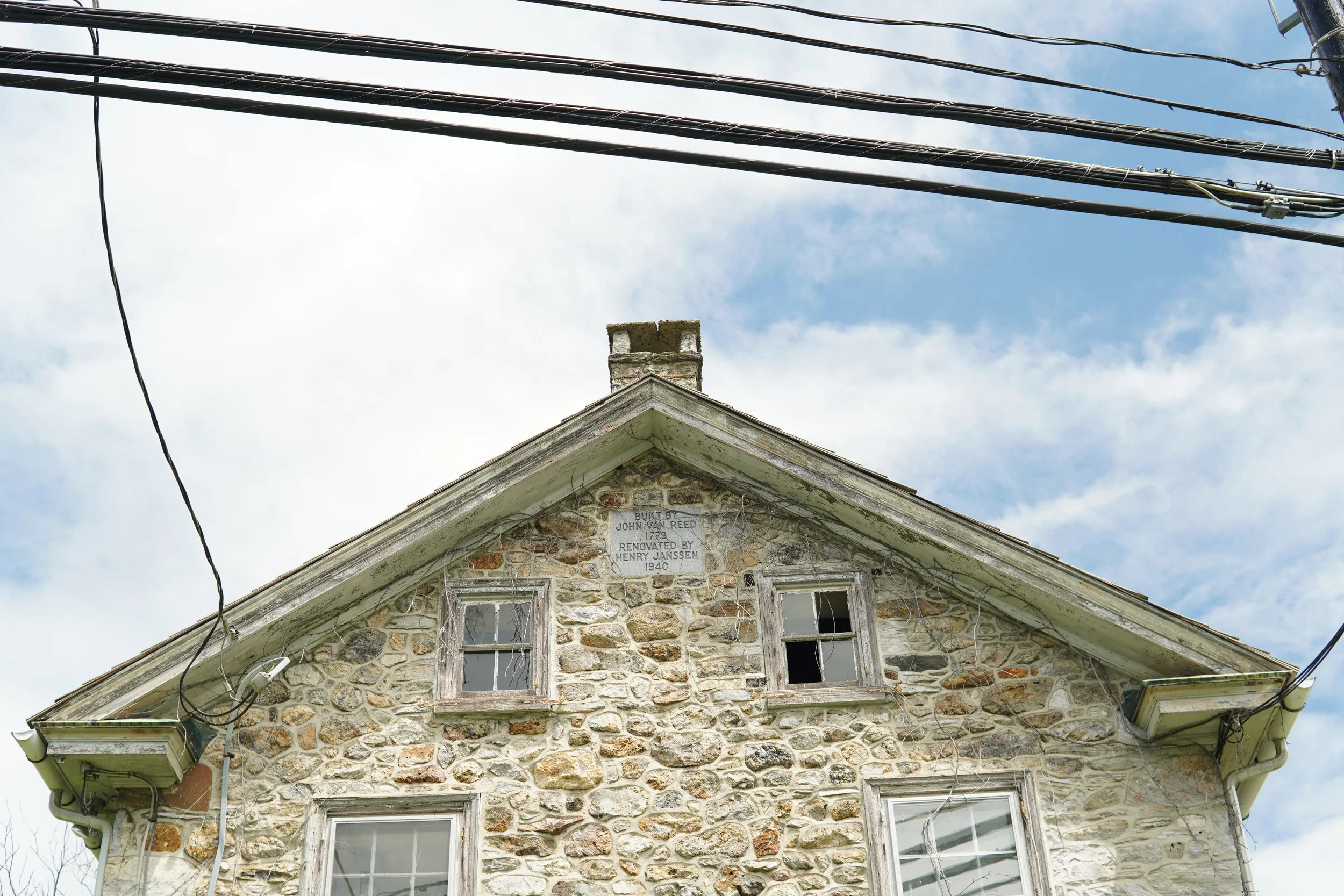 A stone house with a triangular roof that has some windows and a plaque underneath the peak. The sky is partly cloudy, and power lines are overhead.