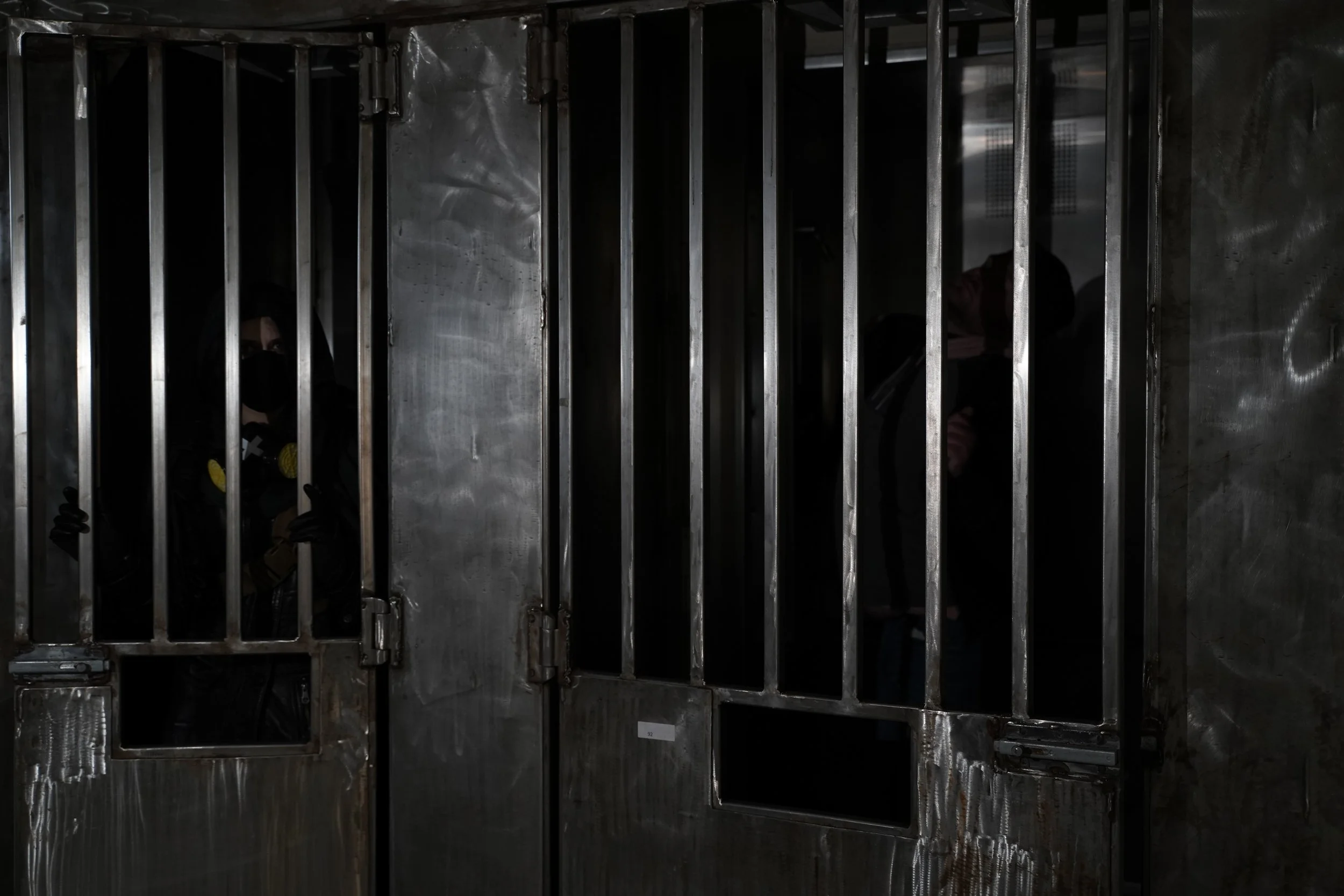 An empty stainless steel jail cell with vertical bars, a door with a lock, and a person lying on the floor behind the bars, partially visible.