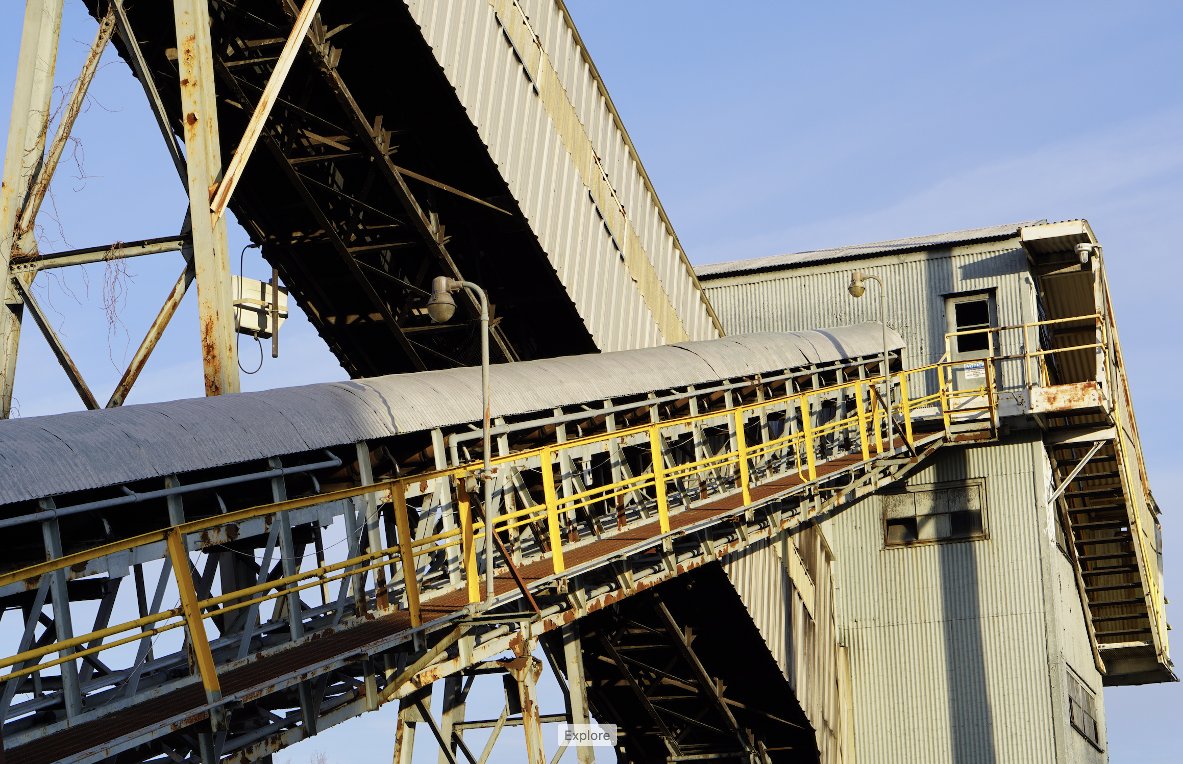 An industrial building made of metal with rusty and weathered surfaces, featuring a conveyor system, stairs, and railings against a clear blue sky.