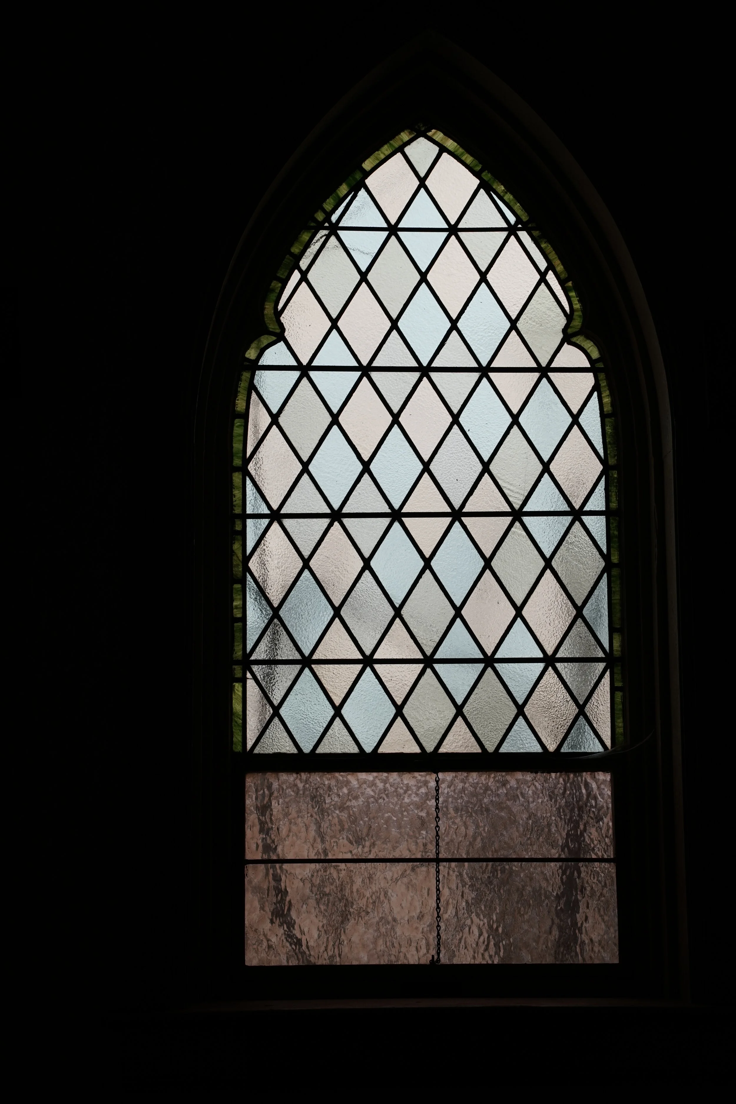 A stained glass window with a diamond pattern at the top and textured glass at the bottom, visible in a dimly lit interior.