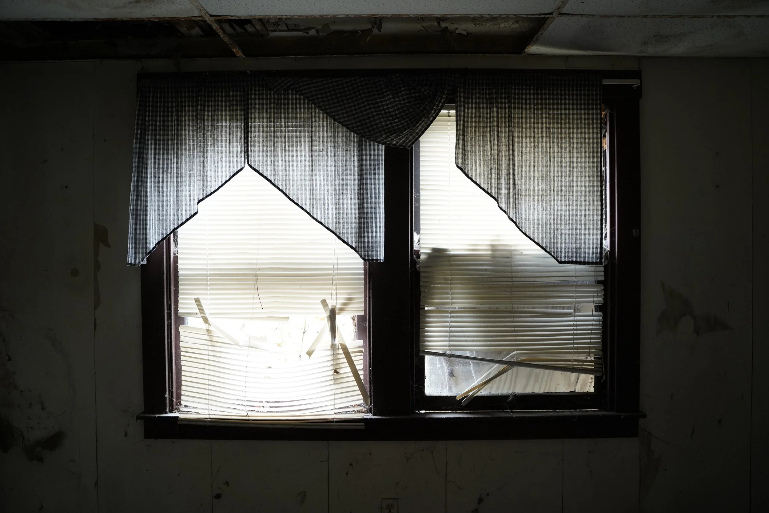 A window with partially broken blinds and tattered curtains in a dilapidated room.