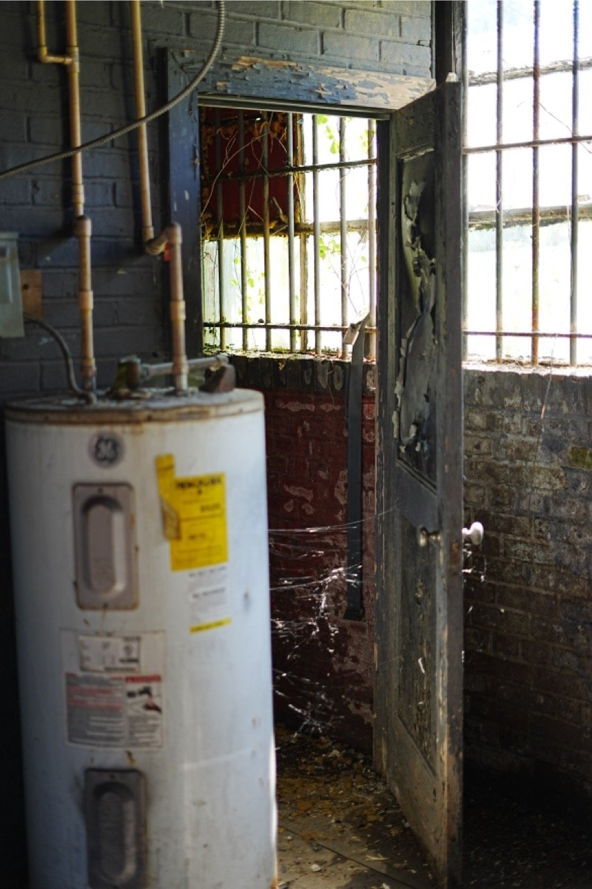 An old, rusty water heater next to a damaged and rusty metal door with spider webs in an abandoned, dilapidated brick building with barred windows, some overgrown with vines.