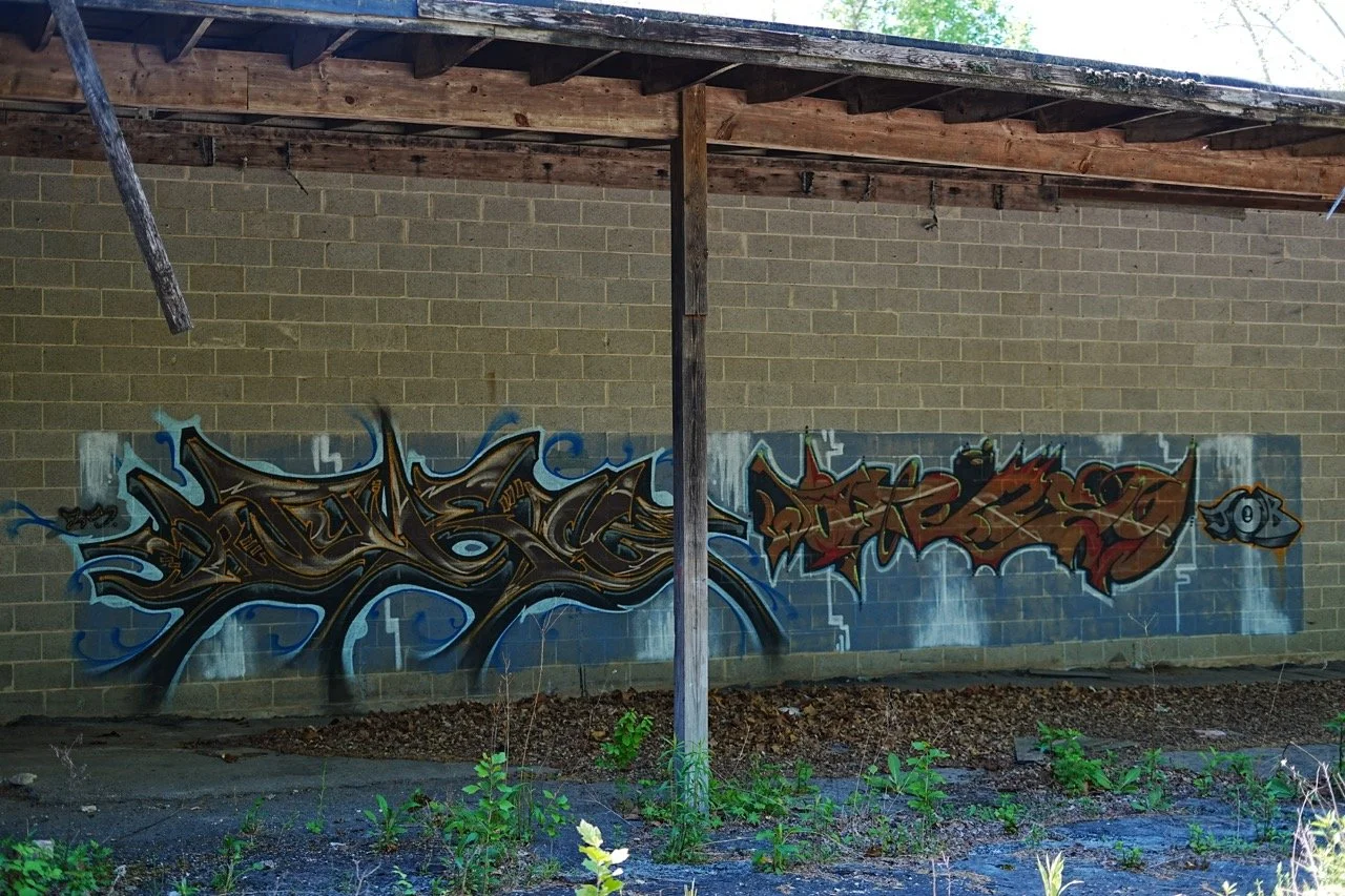 Graffiti art on a brick wall beneath a wooden structure, featuring stylized lettering in shades of black, brown, and red, with blue and white accents.