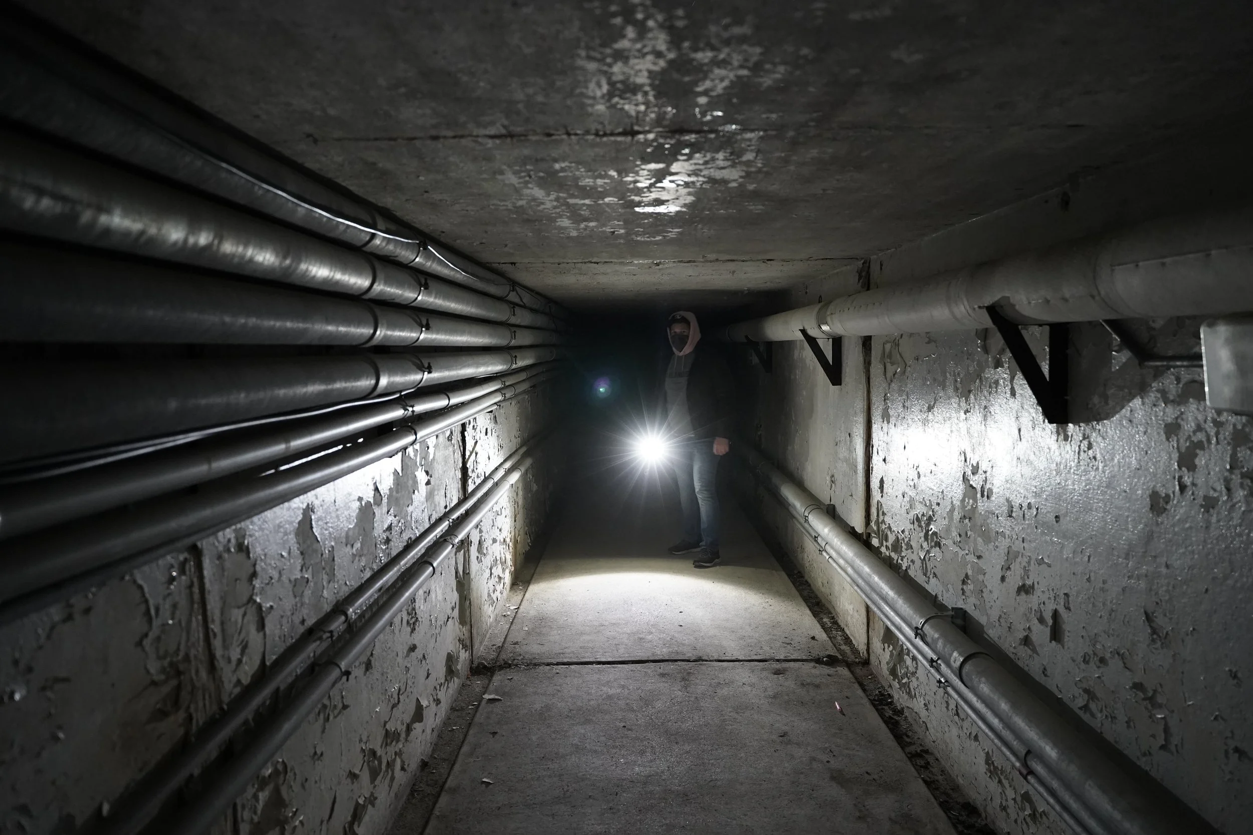A person holding a flashlight walking through an underground tunnel with pipes on both sides and peeling walls.