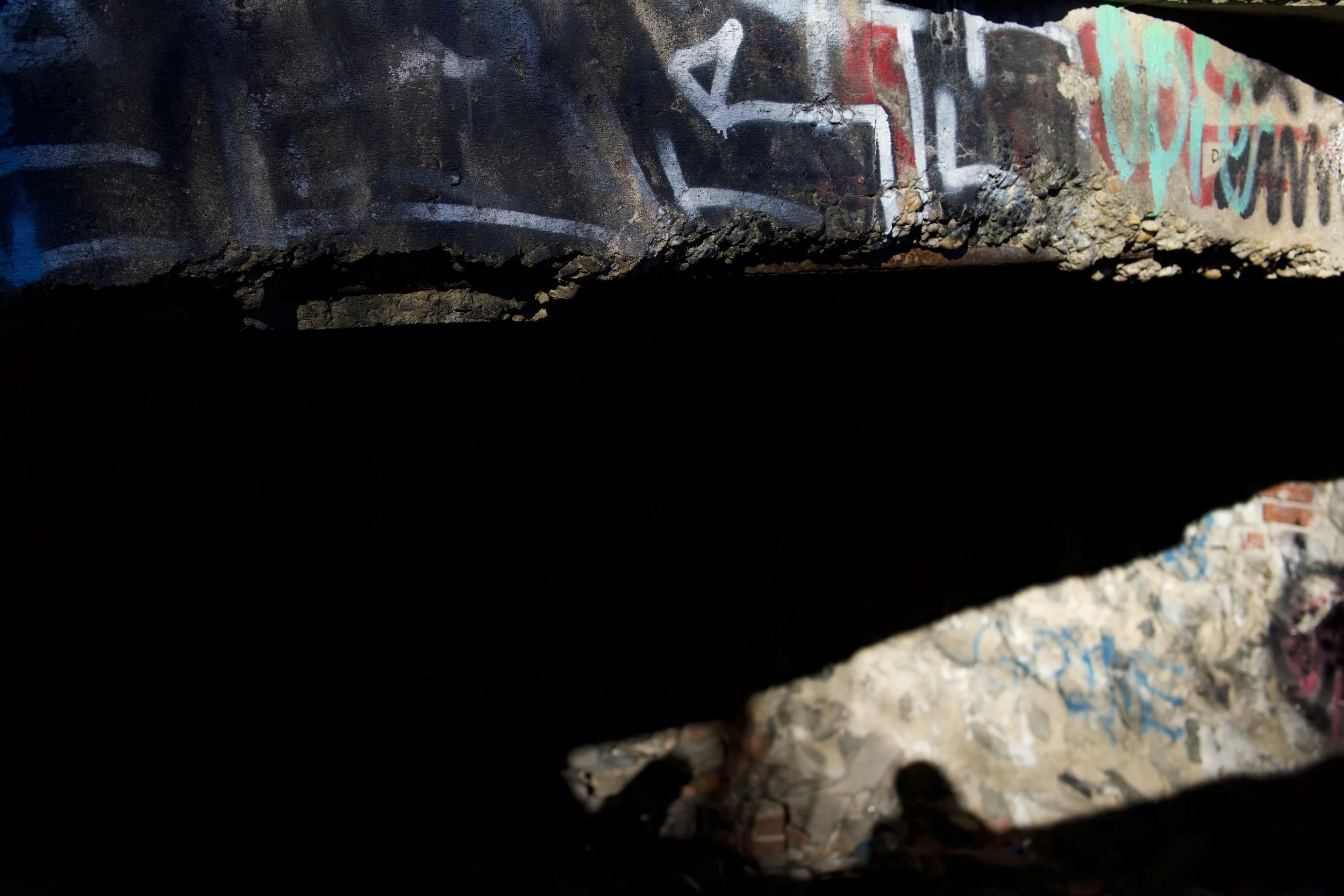 Dark tunnel or crack in the ground with graffiti on the sides, leading to what appears to be an open area with bricks and rubble.