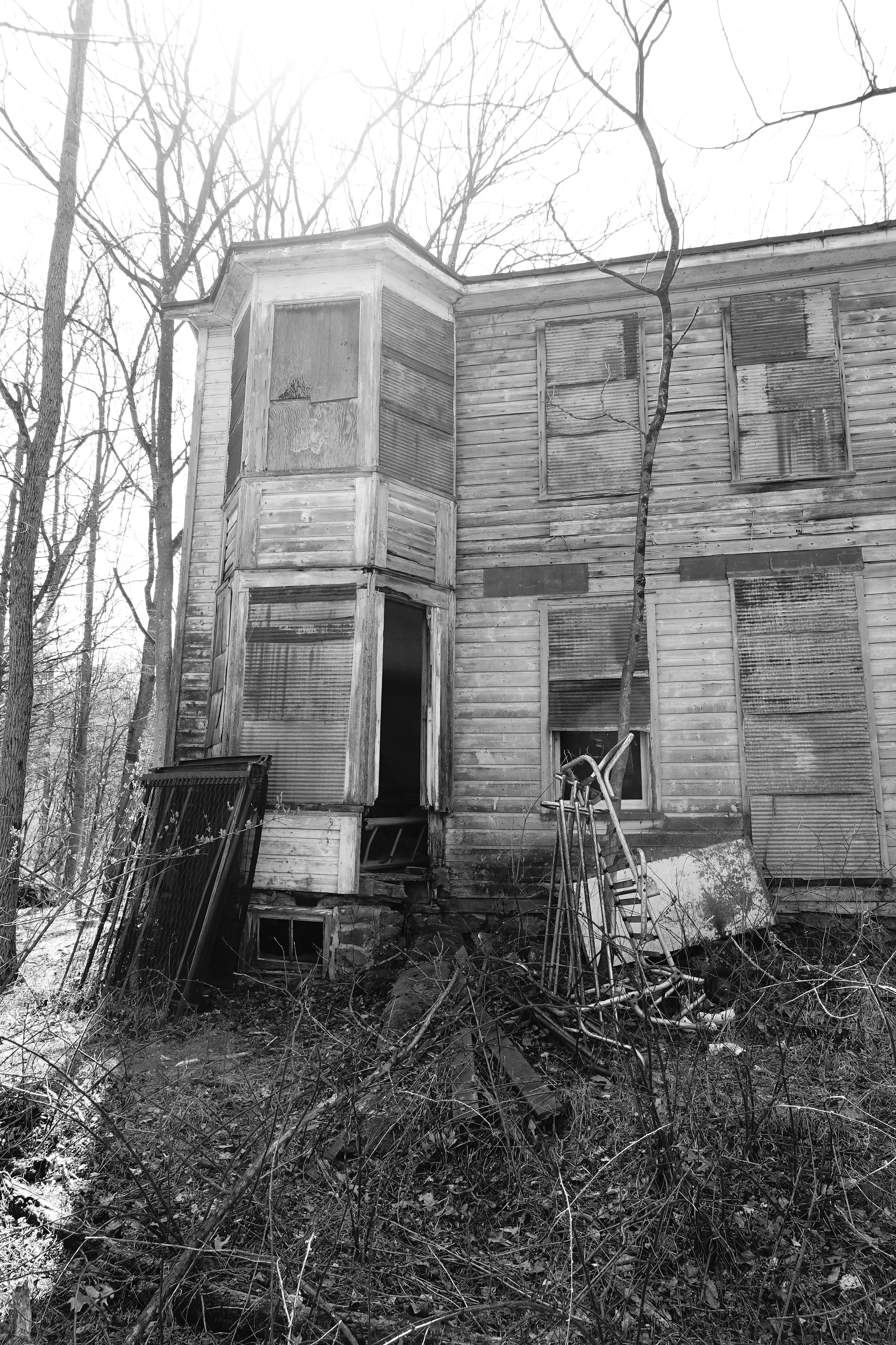 An old, abandoned wooden house with boarded-up windows and a partially open front door, surrounded by overgrown brush and leafless trees on a cloudy day.