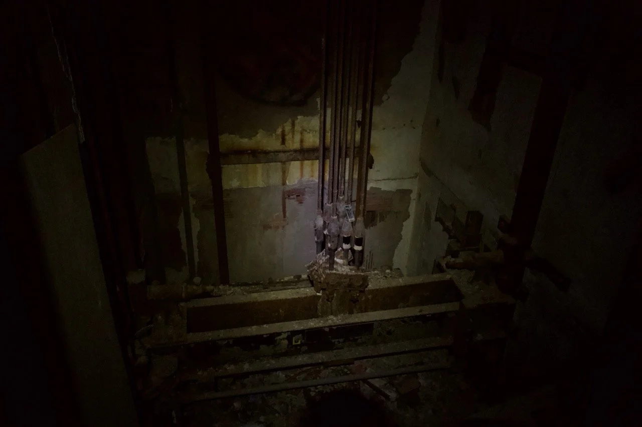 A dark, abandoned elevator shaft with exposed pipes and rusted structural components.