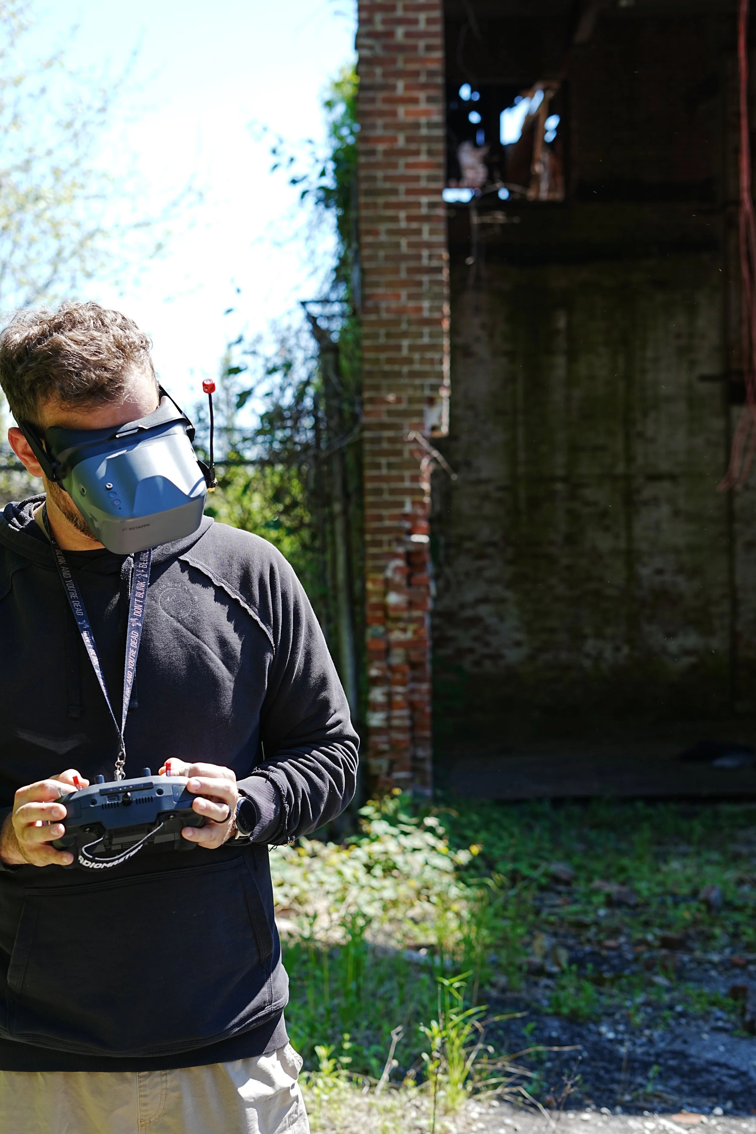 A man with curly hair wearing sunglasses and a virtual reality headset indoors. The background shows a brick wall and a window.