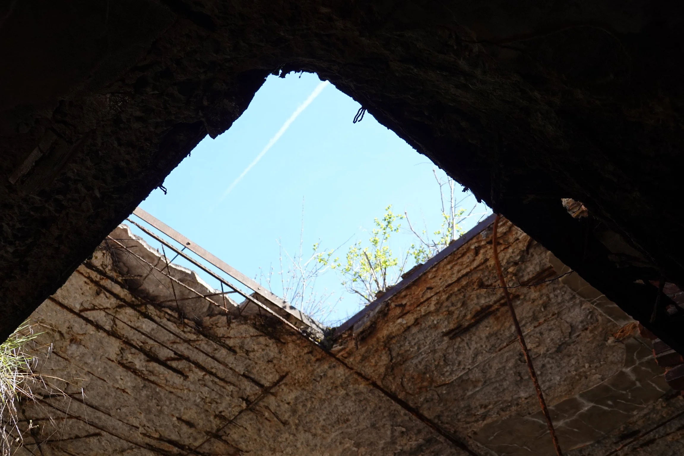 View of the sky through an opening in a dilapidated brick structure, with some metal and vegetation visible.