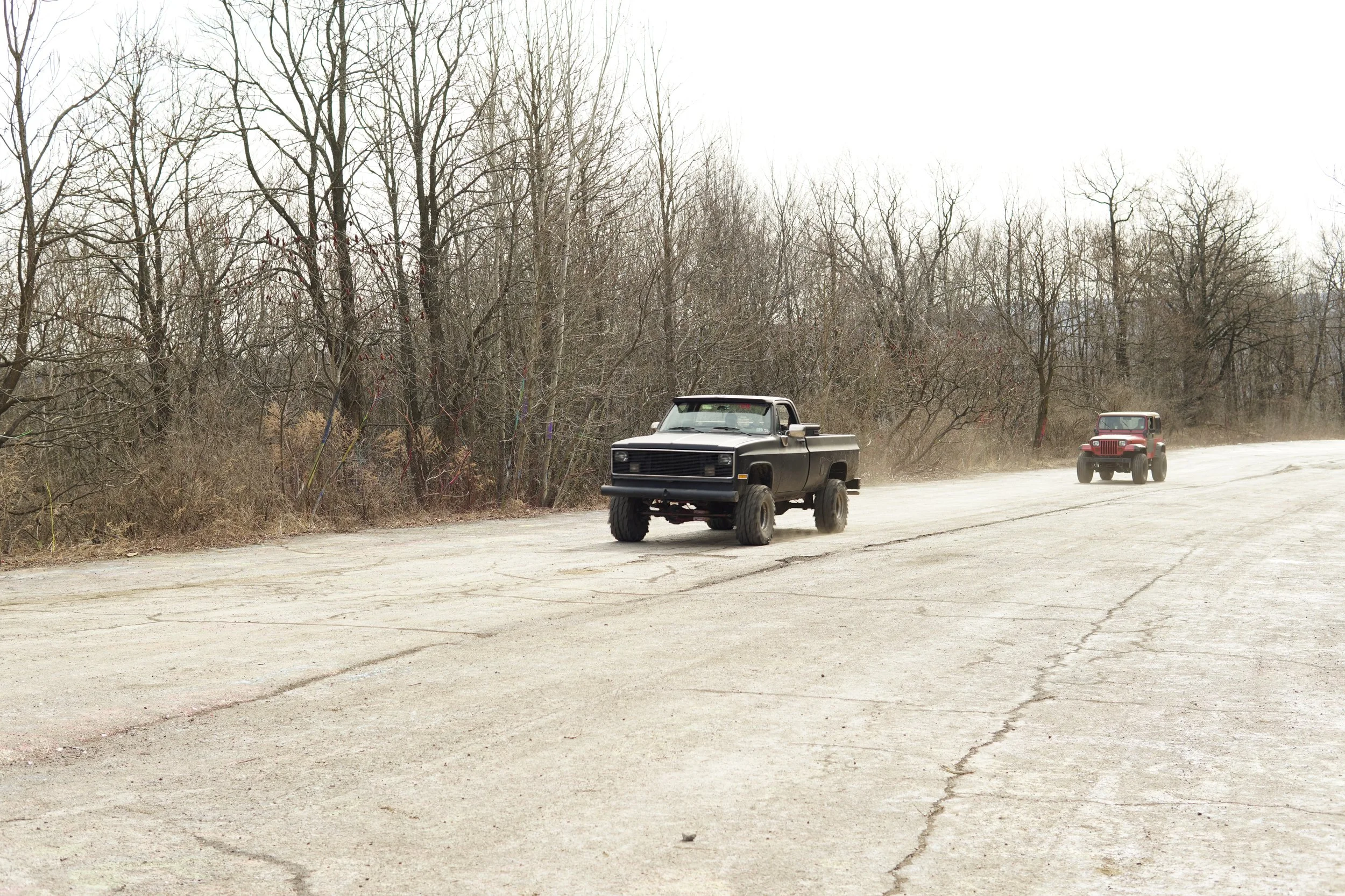 Two vintage trucks, one black and one red, driving on a cracked, dusty road next to leafless trees in a barren landscape.