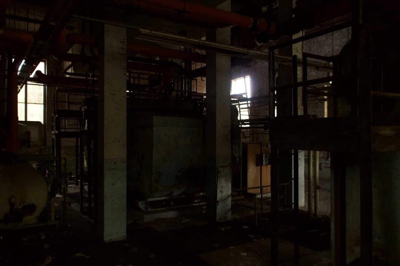 An indoor industrial space with pipes and metal structures, dimly lit with some light coming through a window.