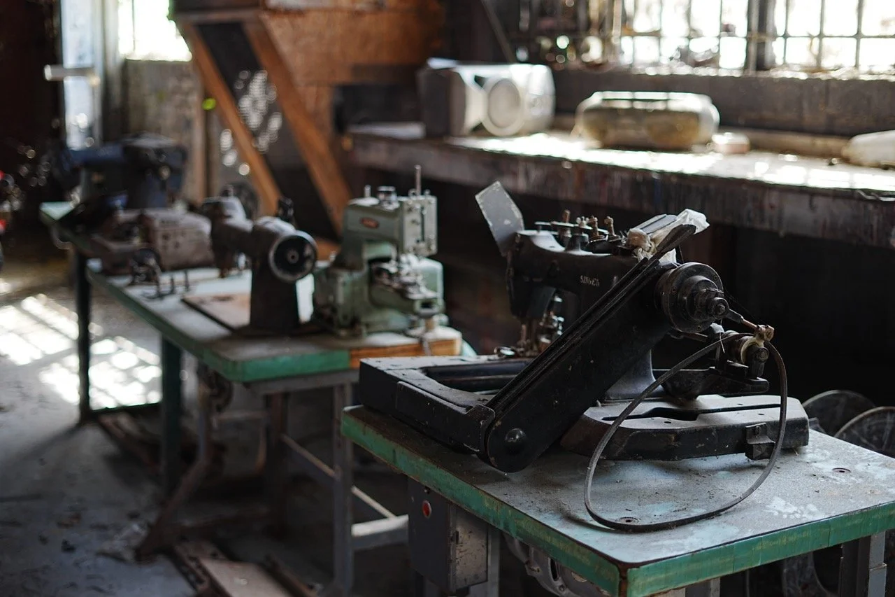 Multiple vintage industrial sewing machines on green tables in an old workshop with wooden walls and windows letting in natural light.