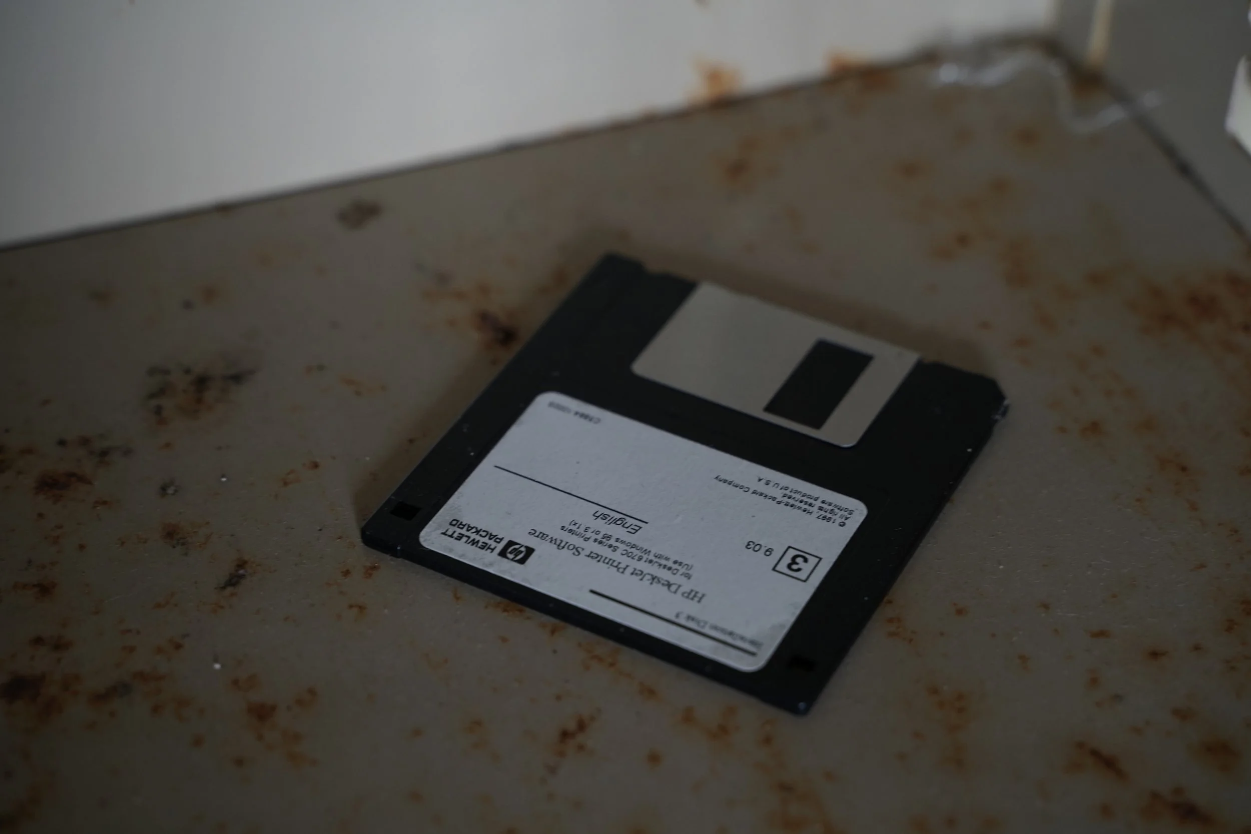 A black floppy disk lying on a speckled surface with a white label, featuring black text and a barcode.