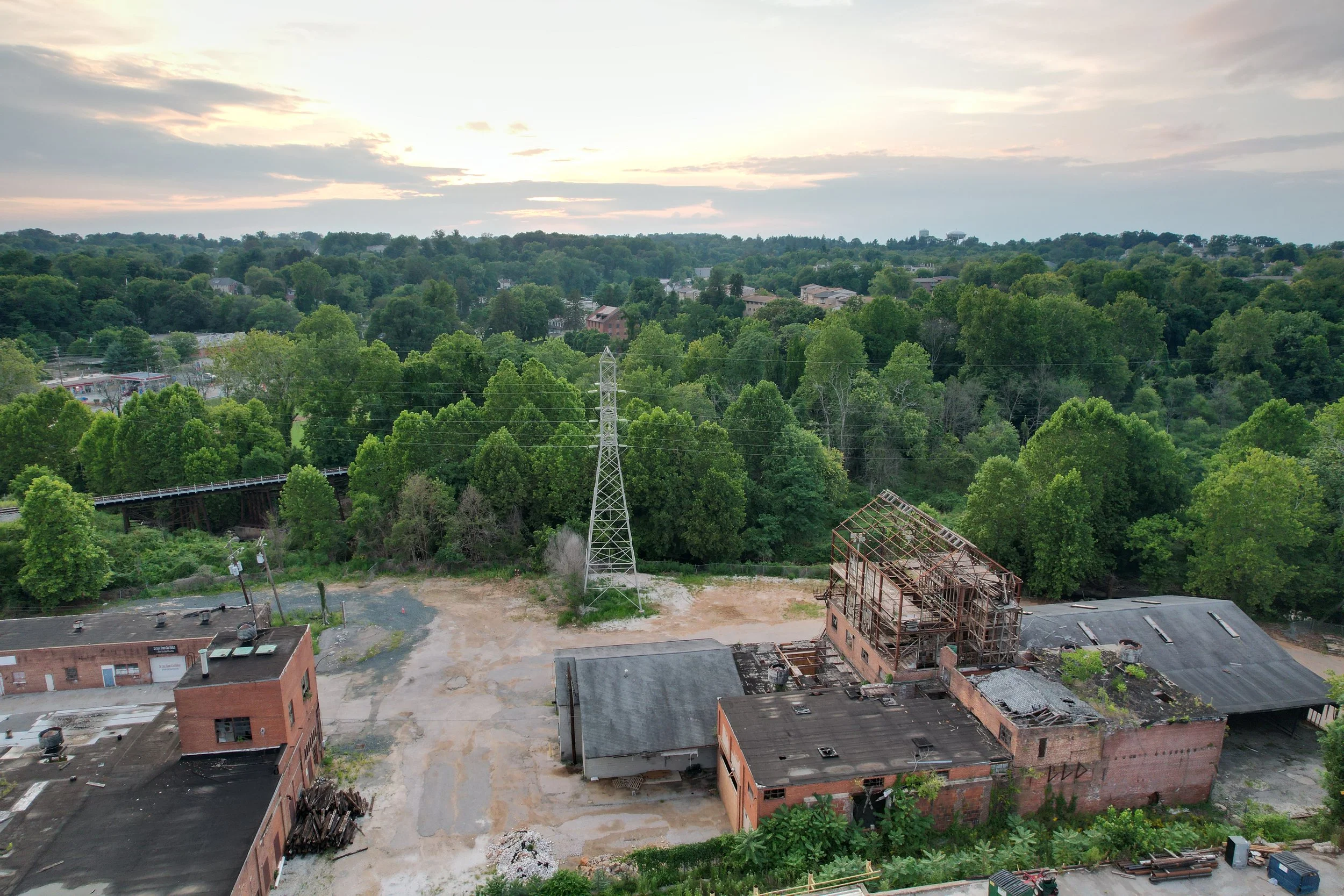 Aerial view of a cityscape with a mix of green trees, residential buildings, and a construction site with an unfinished structure in the foreground, overcast sky.